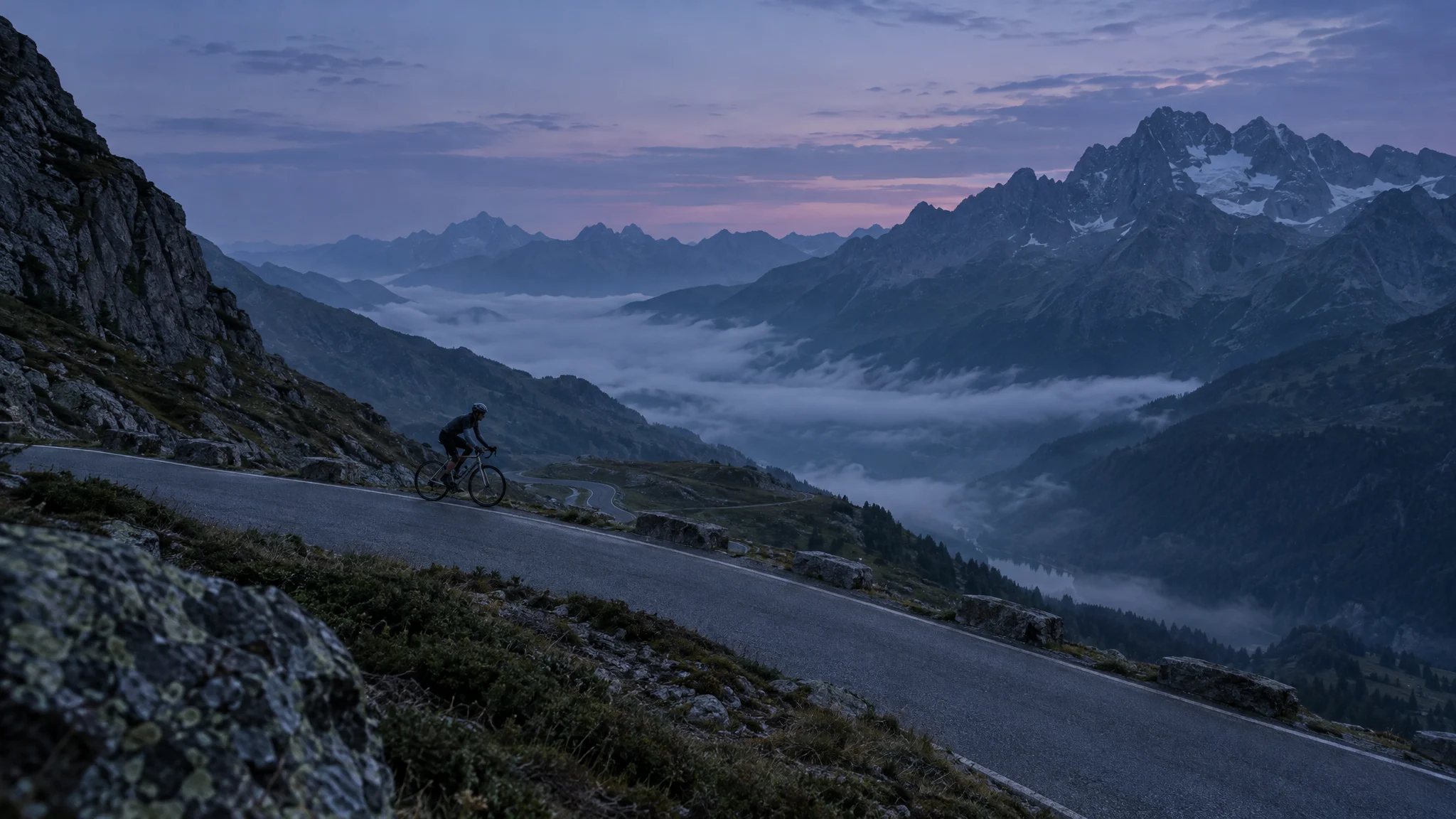 A lone road cyclist climbing a mountain pass at dawn above a valley filled with cloud