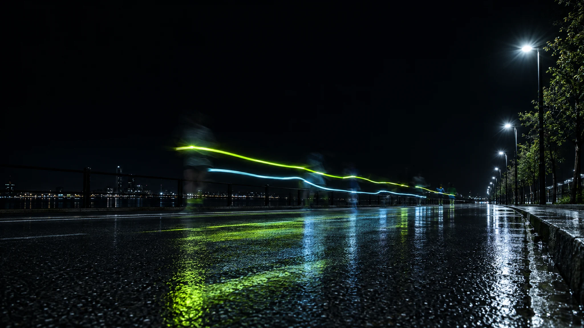 Long exposure of a wet riverside path at night, with lime and cyan light trails left by runners passing under the street lamps