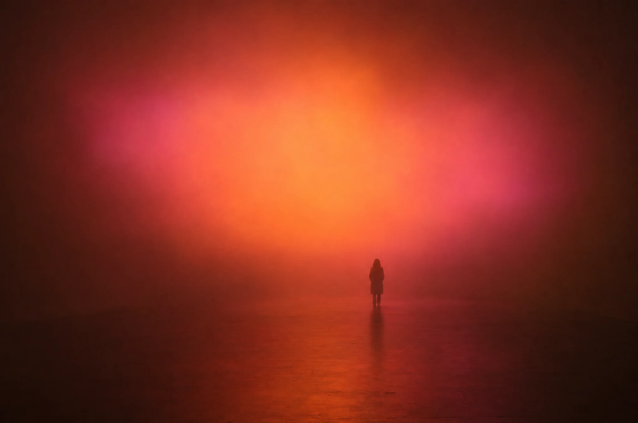 A dark room filled with haze and a wide cloud of apricot, coral and rose light, with one visitor standing at the far end