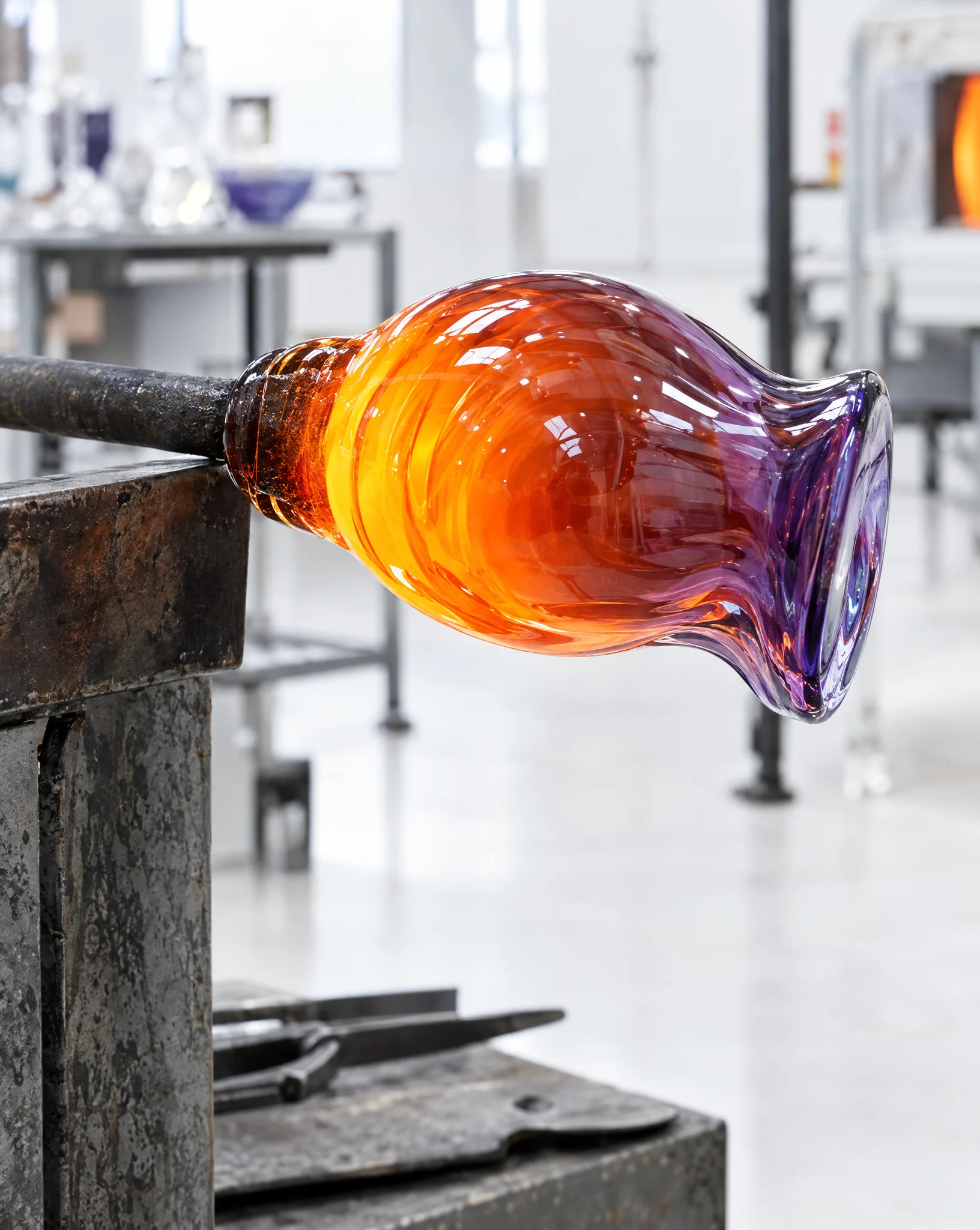 A glowing gather of molten glass, cooling to violet at its lip, held on a steel rod