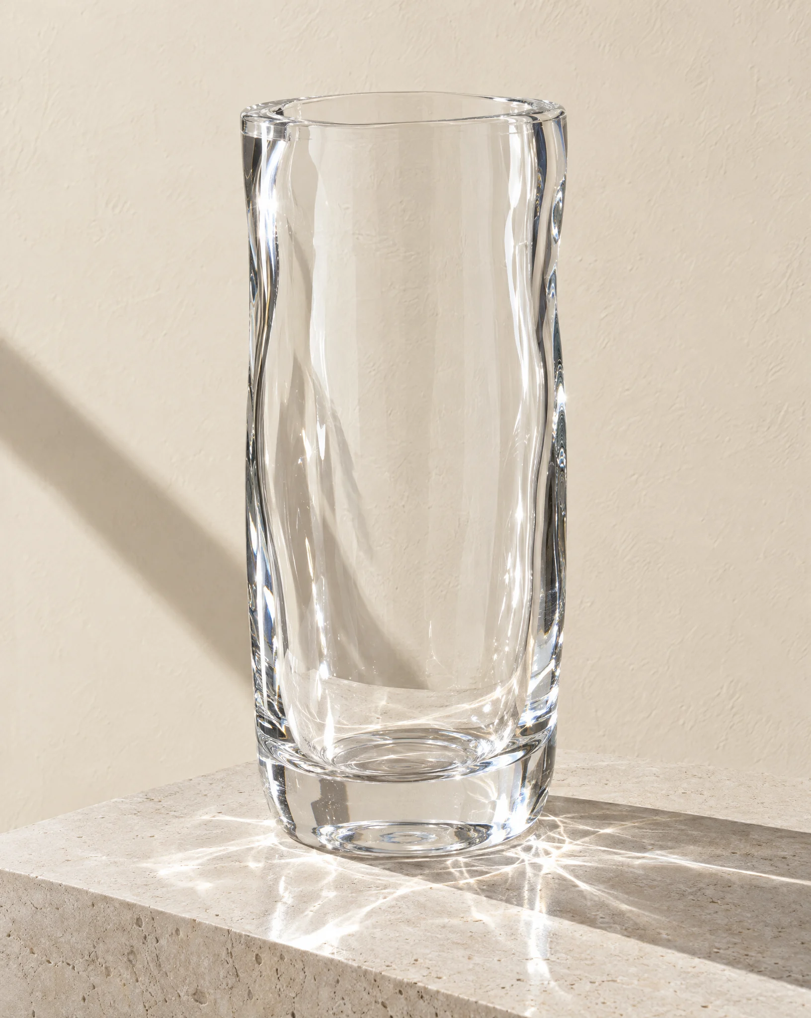 A tall colourless glass vase with softly rippled walls throwing caustic light onto stone