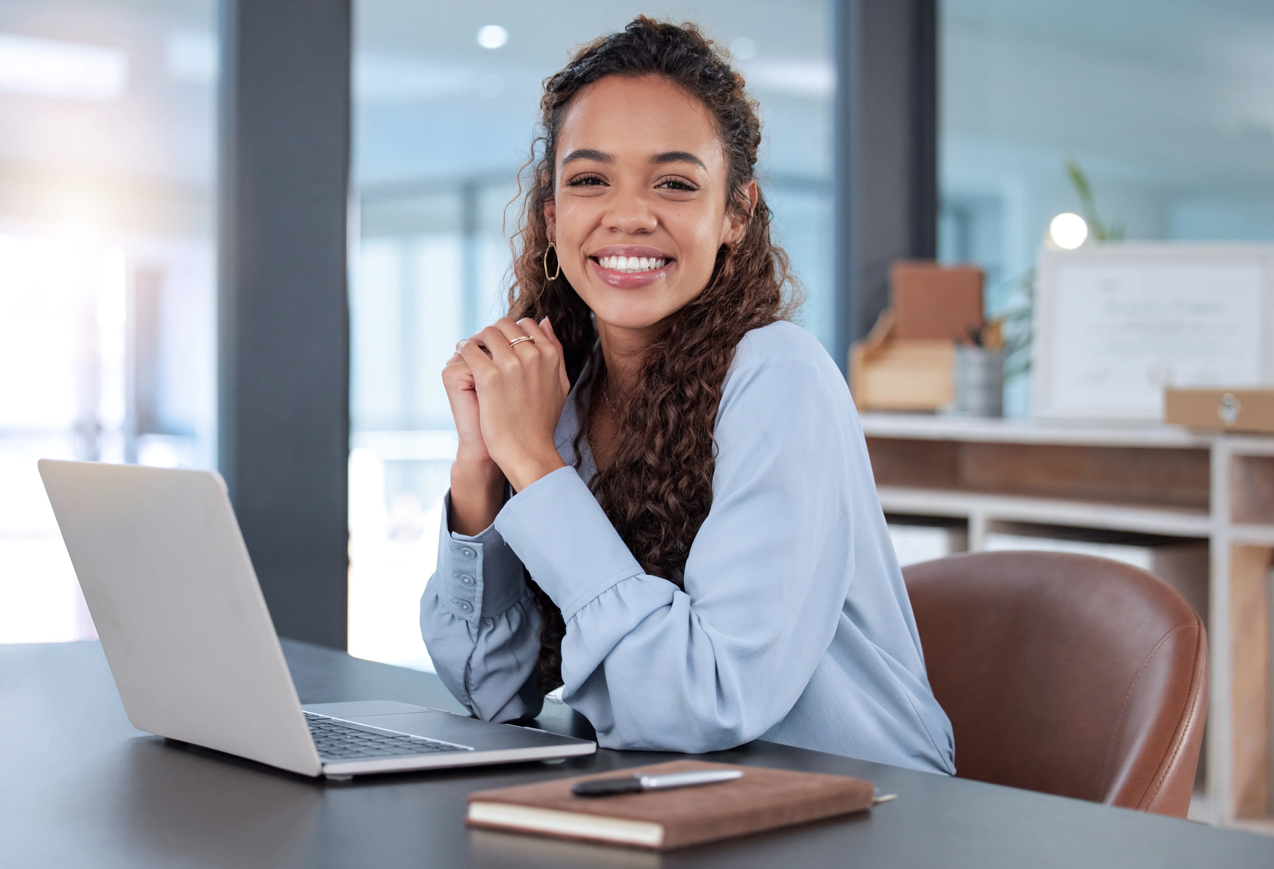 A smiling woman seated at a desk with a laptop and notebook