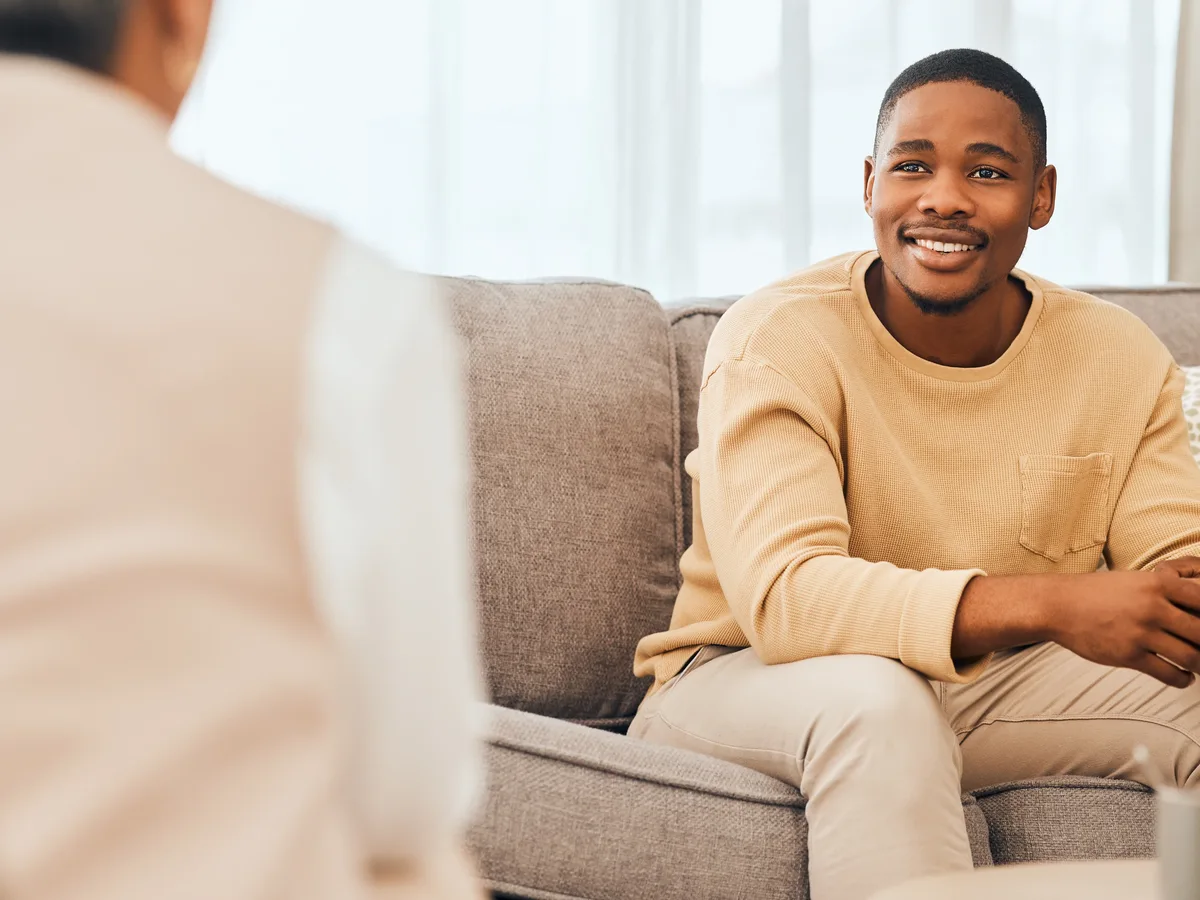 A smiling man seated on a sofa, speaking with someone opposite him