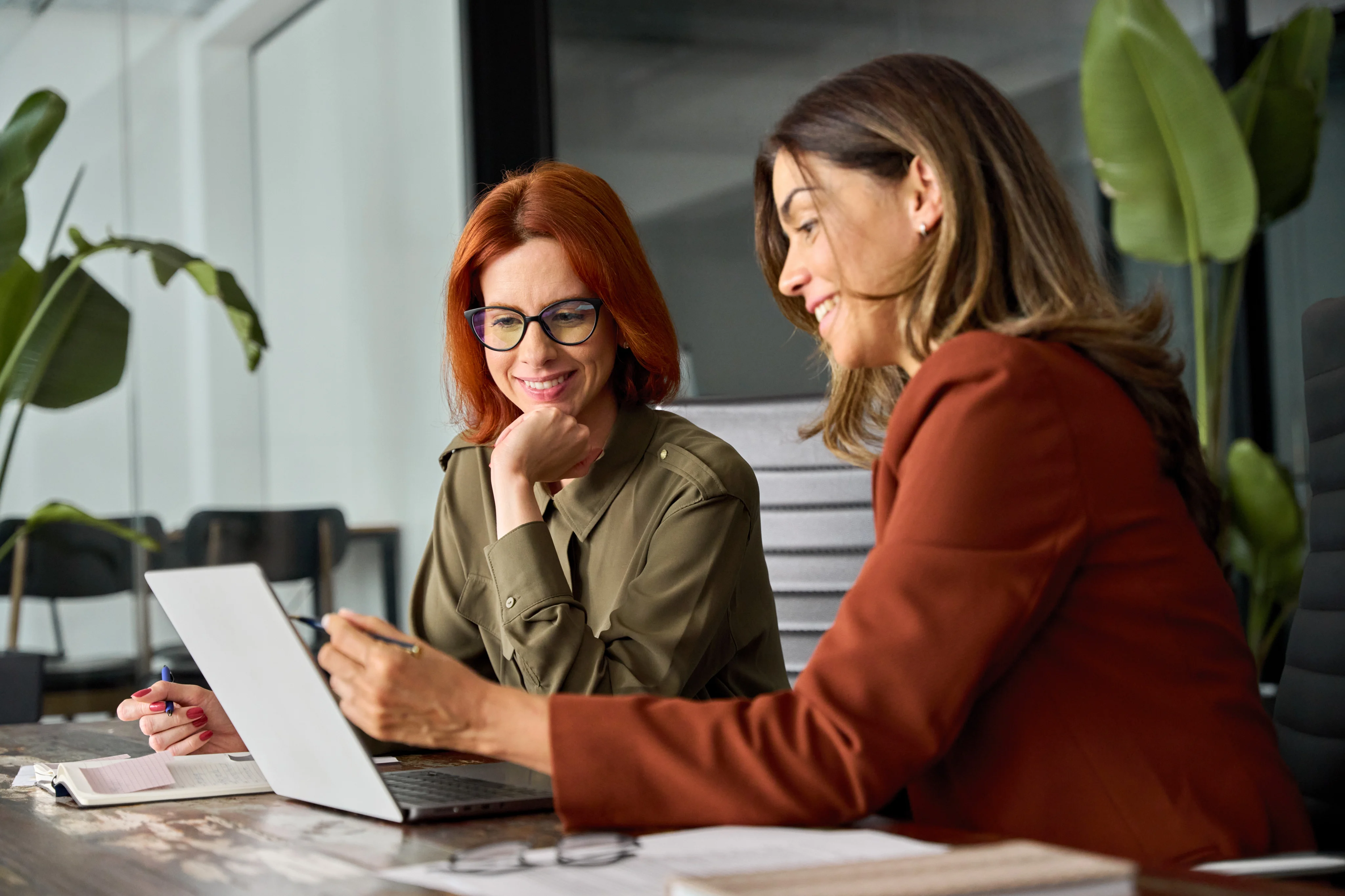Two women smiling while reviewing a laptop together in an office