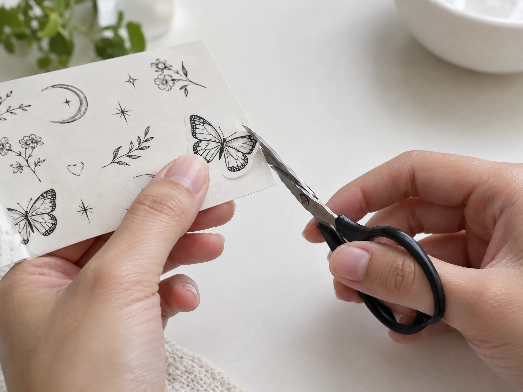 Hands trimming a fine-line butterfly temporary tattoo from its sheet with small scissors