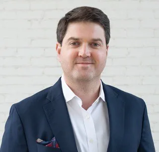 Brent Vegliacich, a confident man in a navy suit and crisp white shirt, stands against a bright white brick wall.