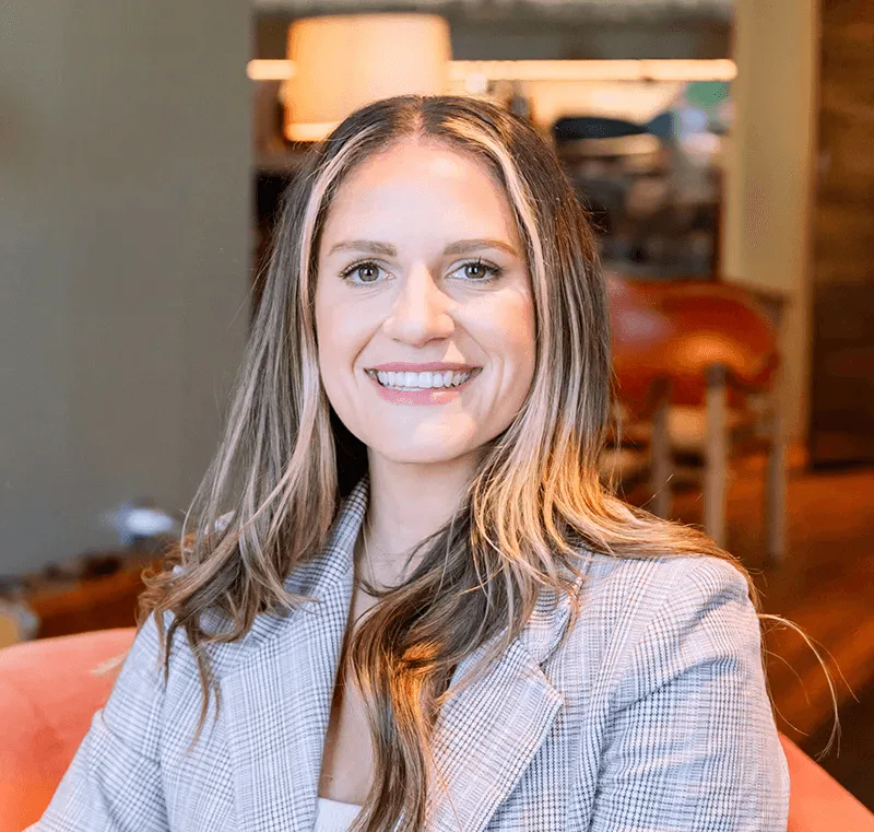 Kaitlyn O'Connor, a smiling woman with long brown hair and blonde highlights, wearing a grey plaid blazer representing a.