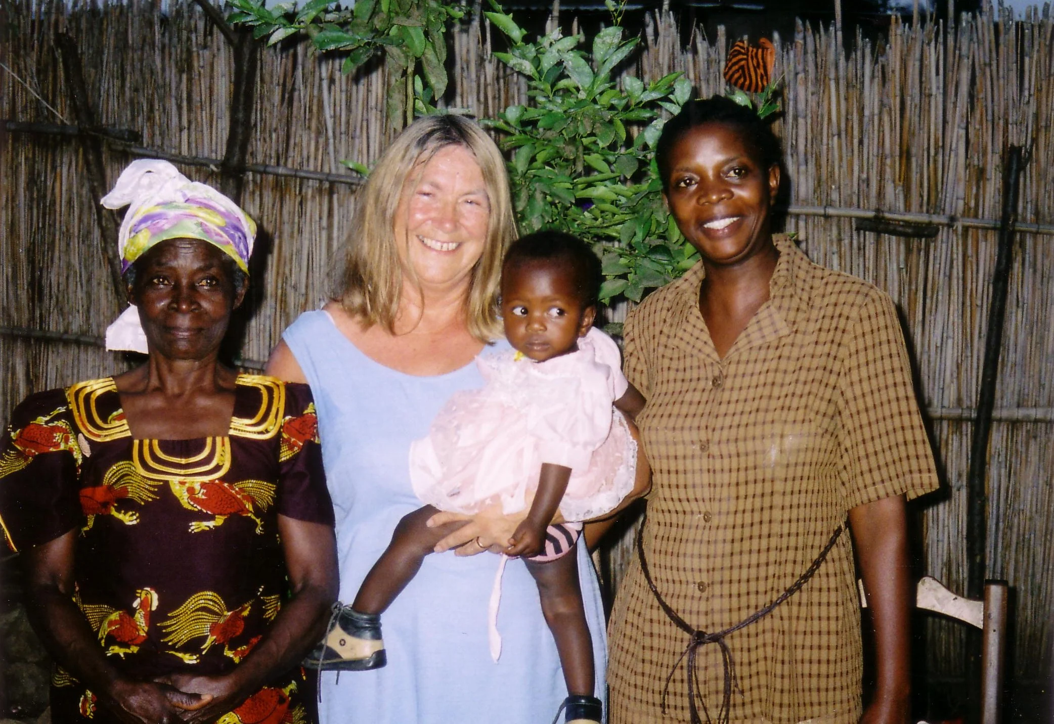 Elaine Storkey smiling beside two women, one holding a young child, in front of a reed fence