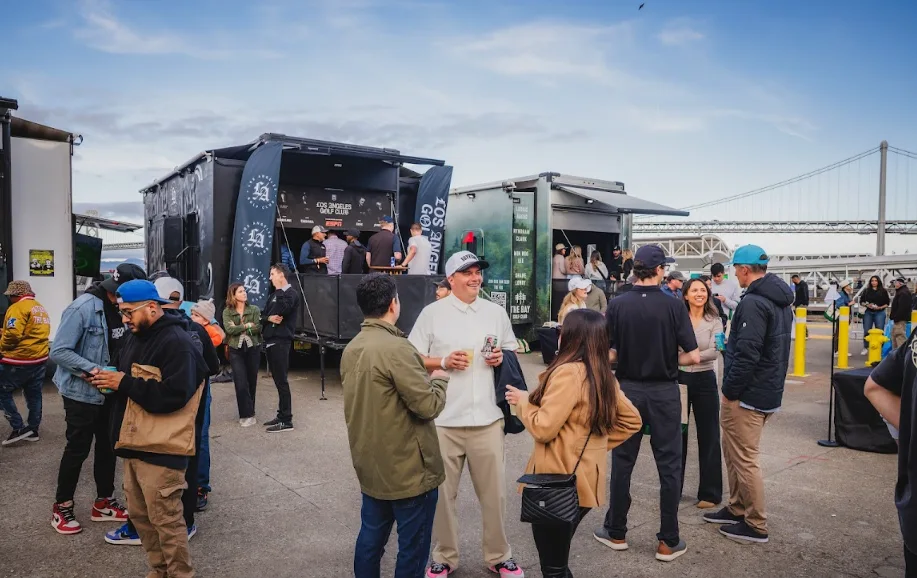 Guests with drinks hanging out beside Dryvebox trailers while others play from the party deck at a waterfront event