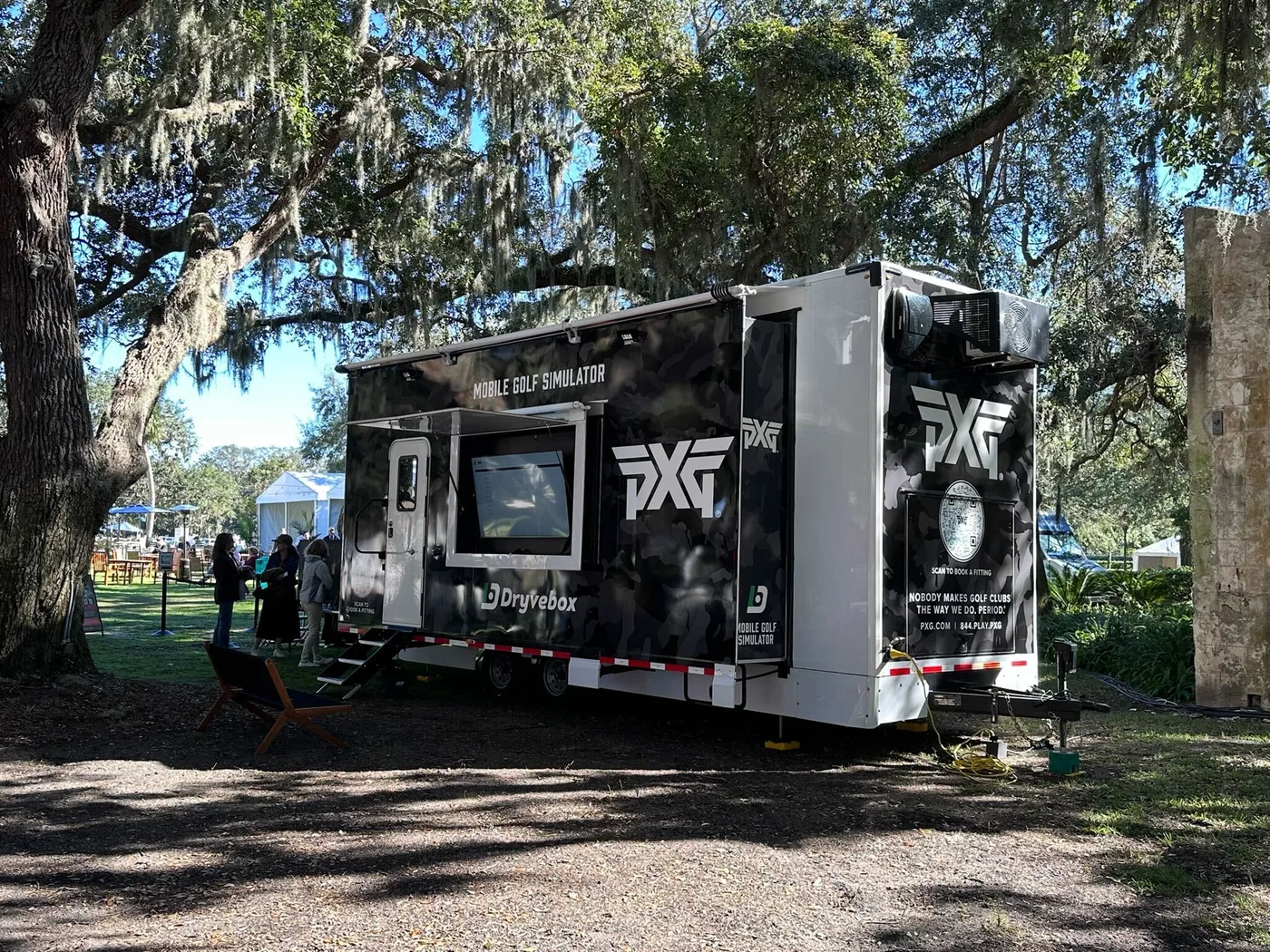 PXG-wrapped Dryvebox mobile golf simulator trailer parked under live oaks on tournament grounds