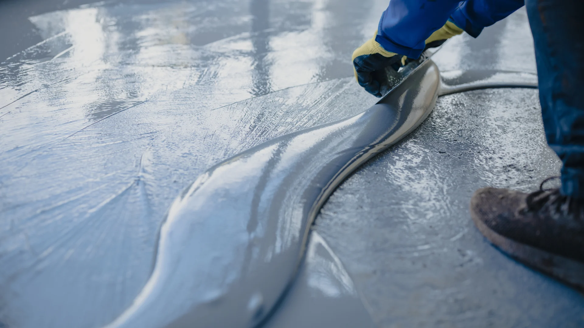 A worker wearing gloves spreads wet gray epoxy resin across a floor using a manual squeegee tool.