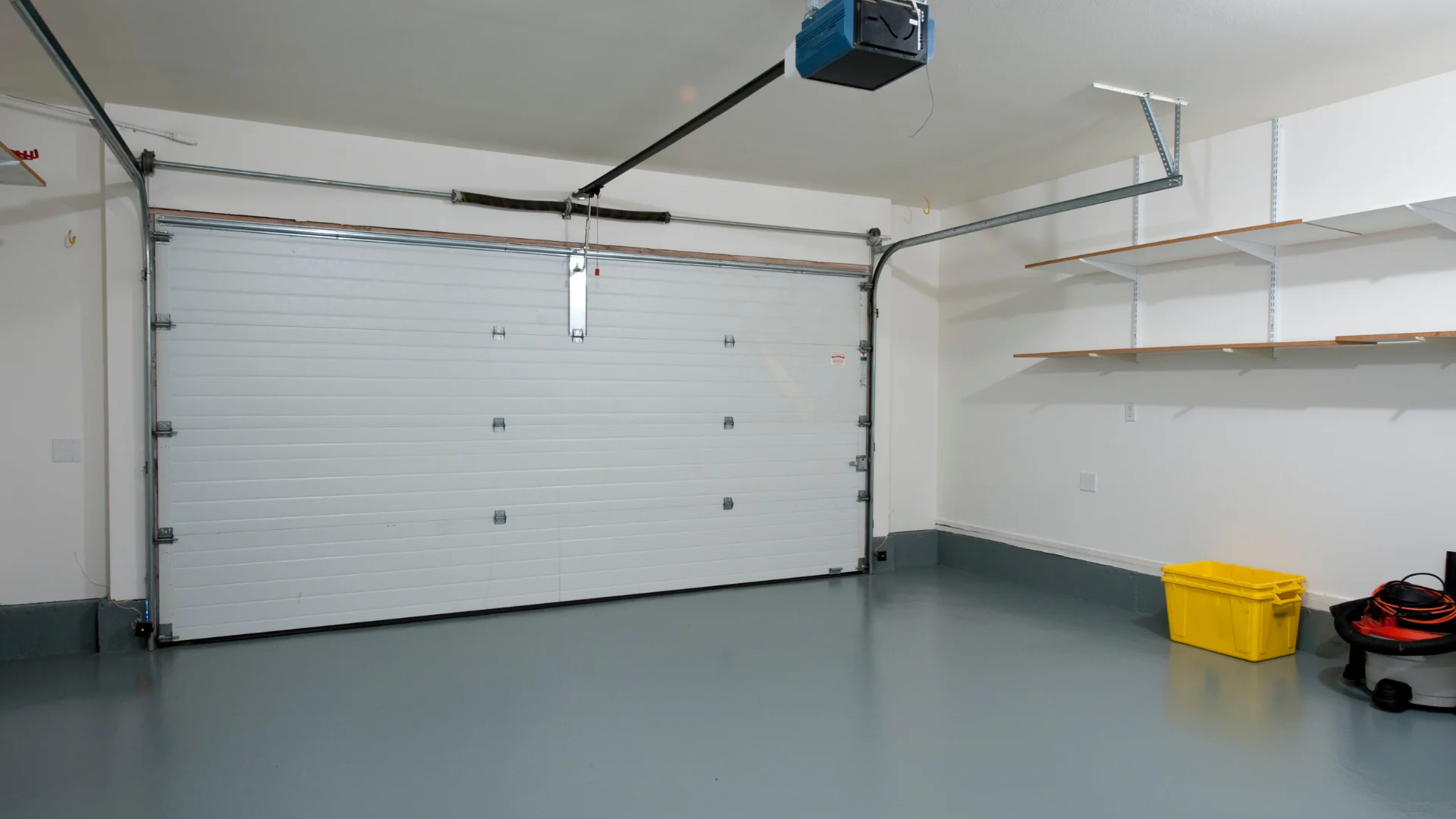 A clean, white, residential garage with a closed sectional door, grey floor, wall shelving, and a yellow plastic bin.