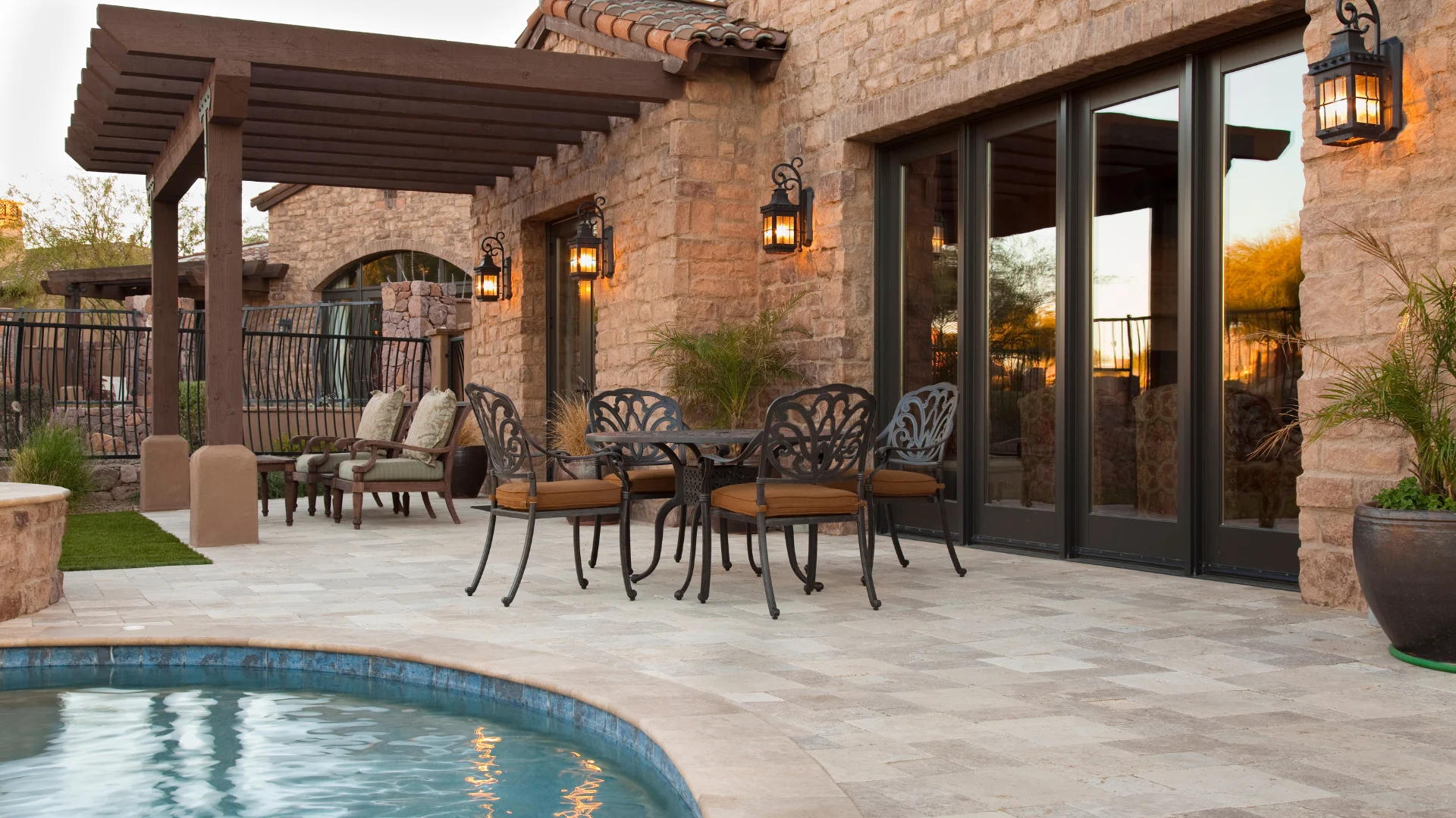A stone patio with a dining set and lounge chairs beside a swimming pool, adjacent to a rustic house with a pergola.