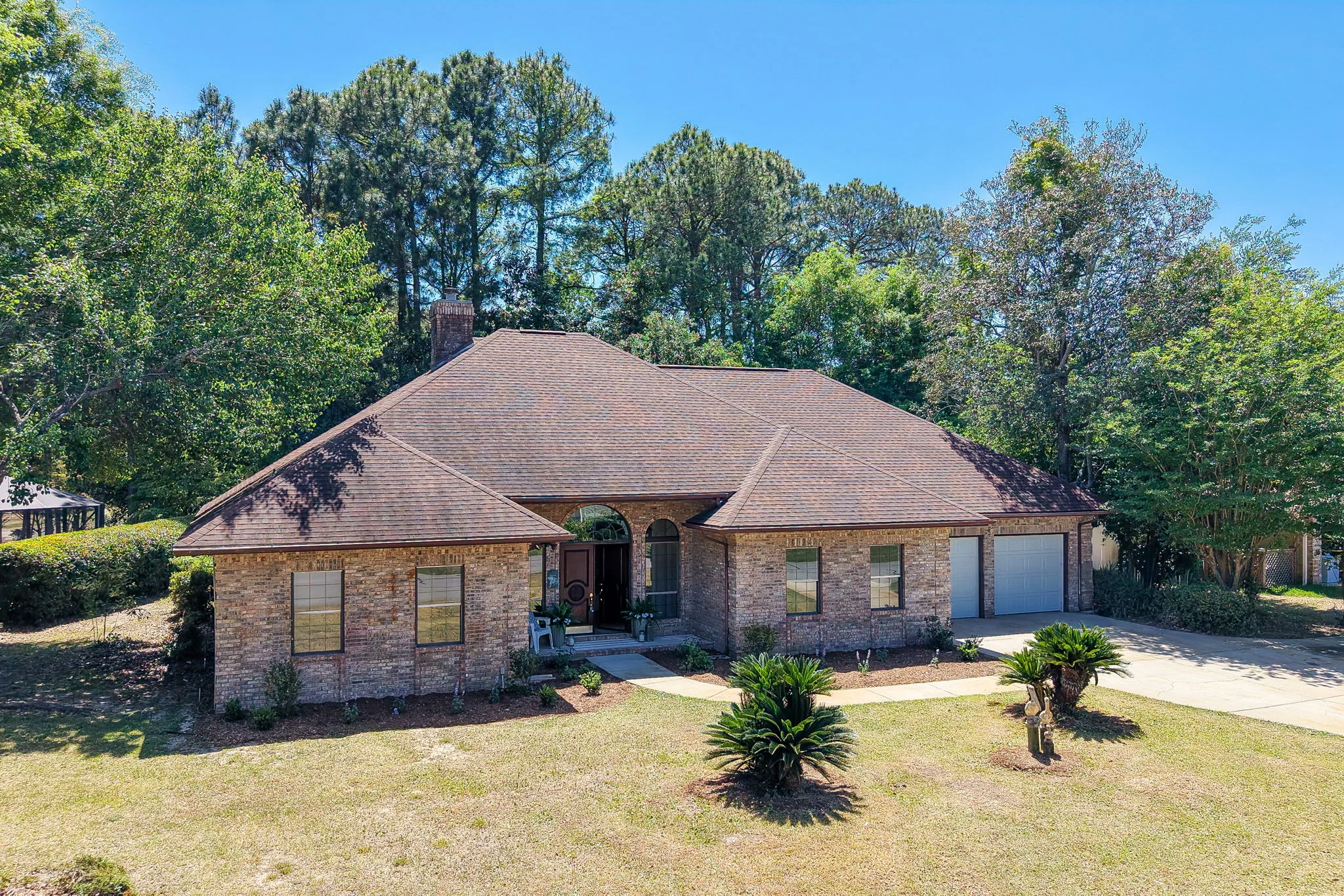 Single-story brick house with a broad shingled roof, garage, lawn, palms, and surrounding trees