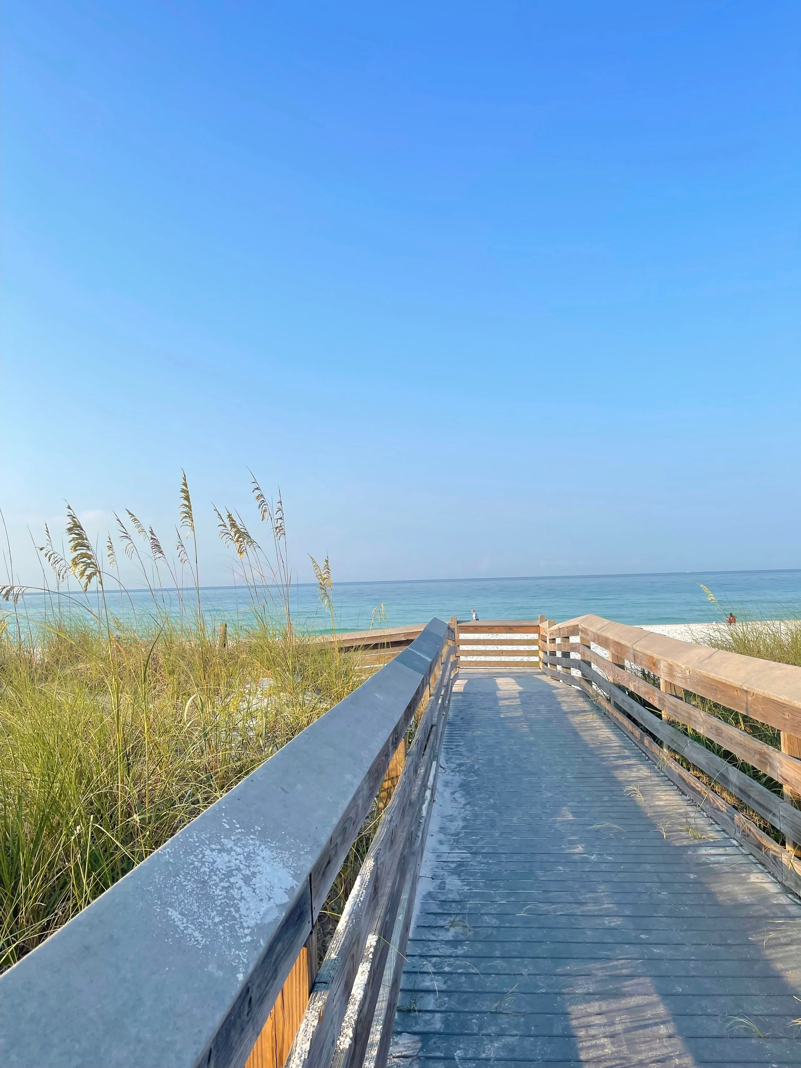 Wooden boardwalk through beach grasses leads toward calm blue water under a bright sky.