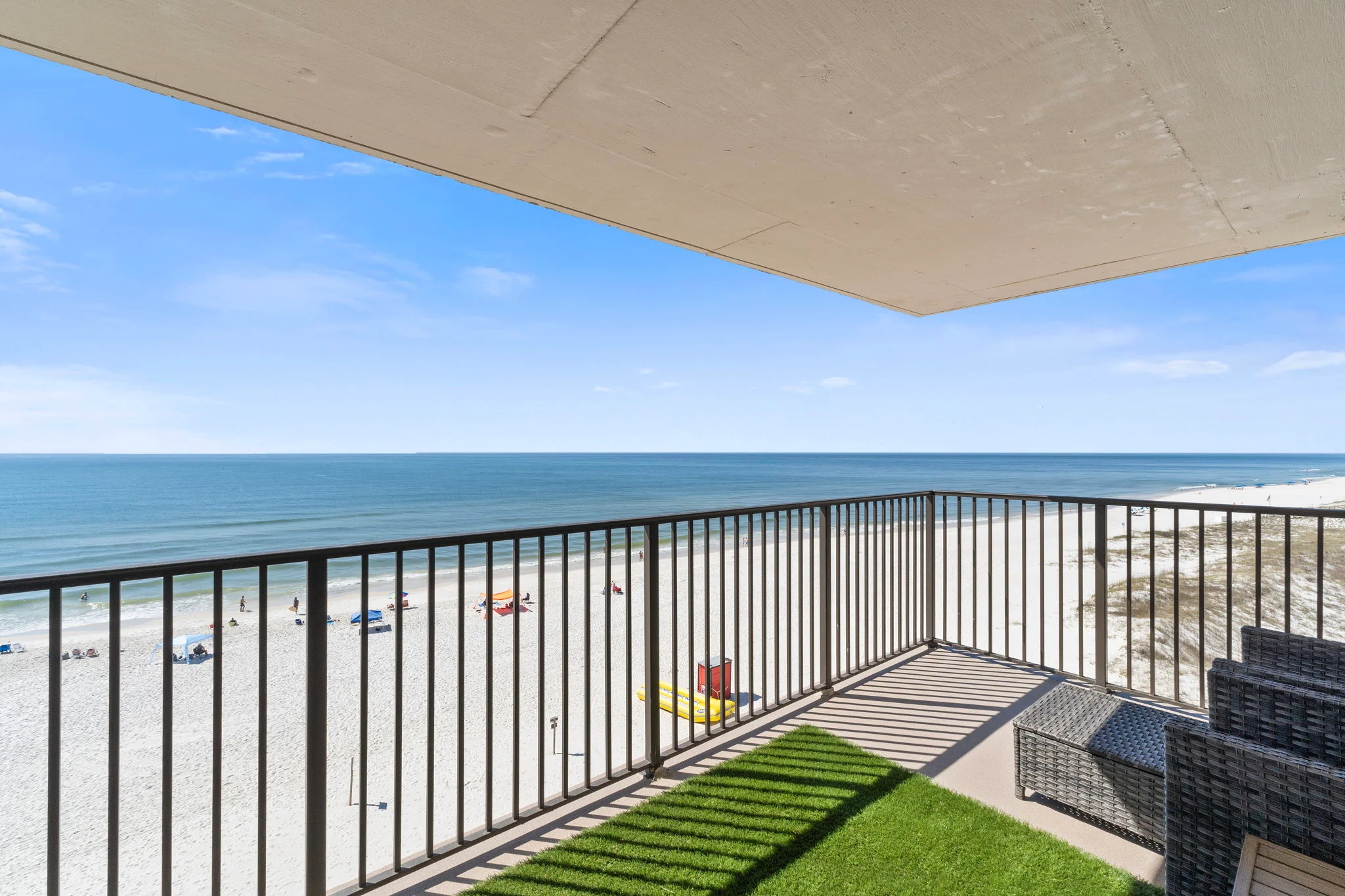 Sunny balcony with dark railings overlooking a white-sand beach, calm ocean, and scattered beachgoers.
