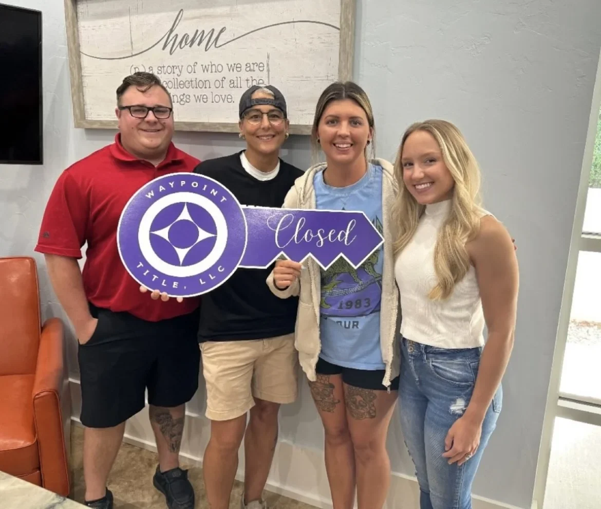 Smiling buyers and the Sumner Realty team hold a large purple Closed key sign on closing day.