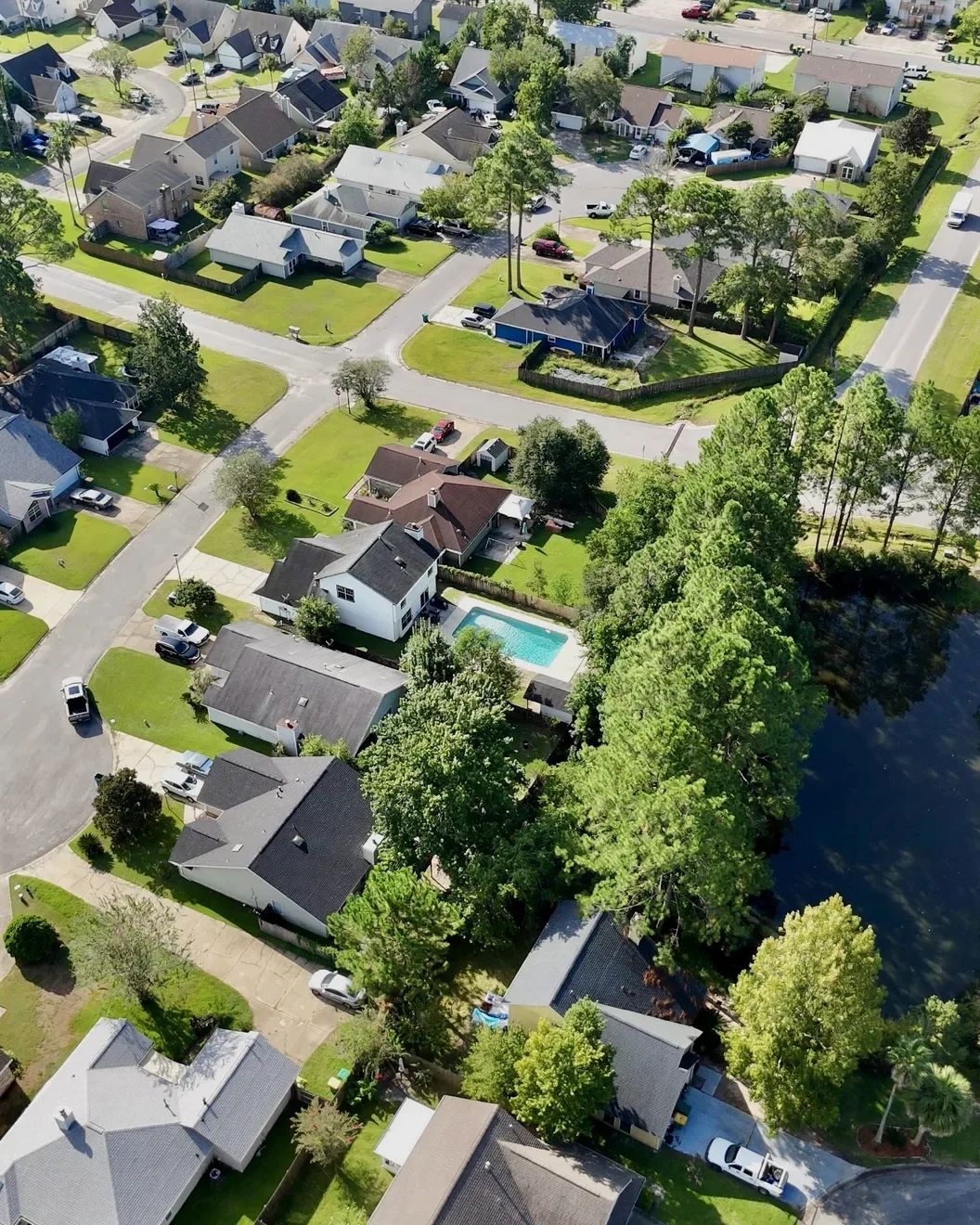 Aerial view of a tree-lined neighborhood with a backyard pool beside a wooded pond.