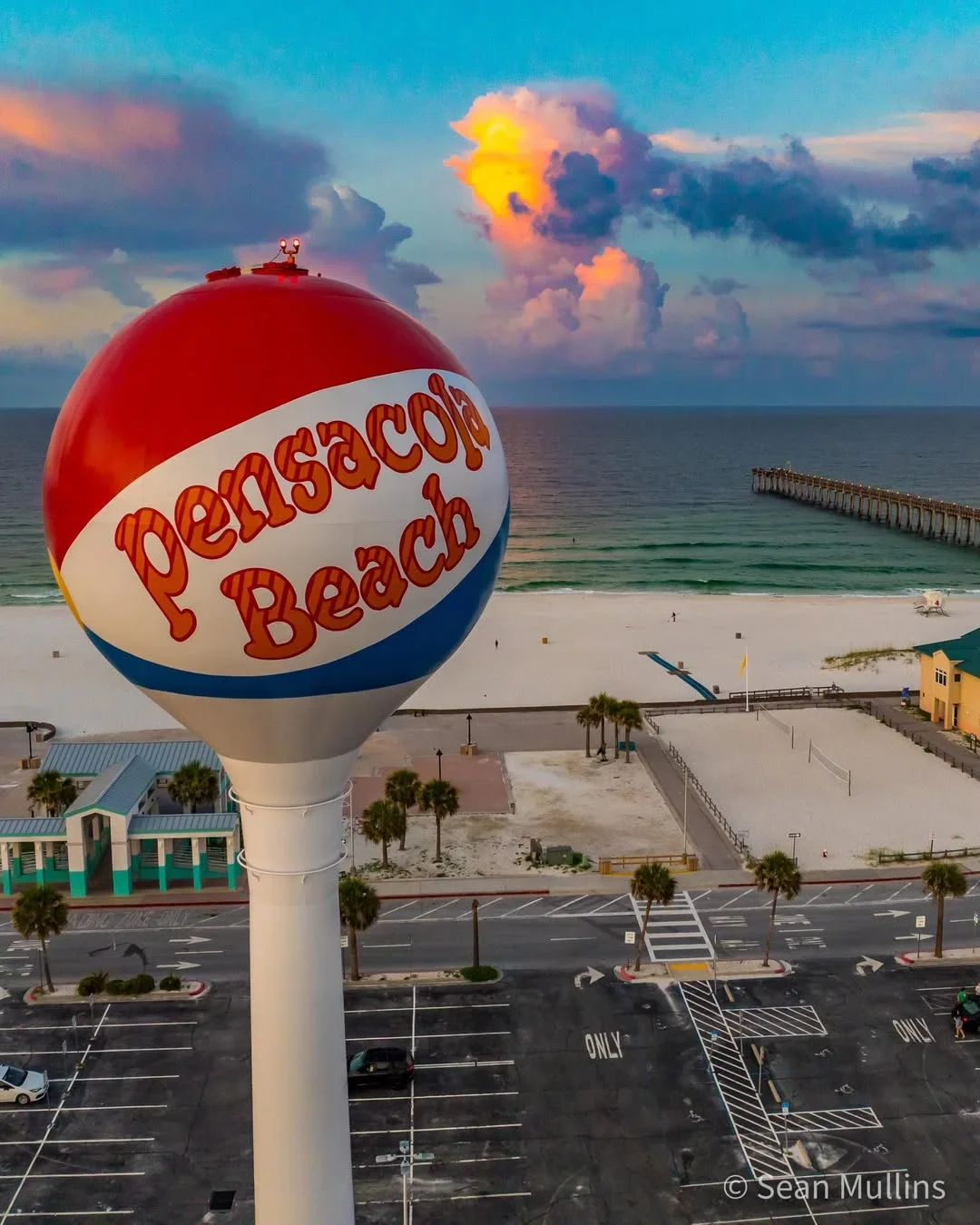 The Pensacola Beach ball water tower above white sand, the Gulf and a fishing pier at sunset.