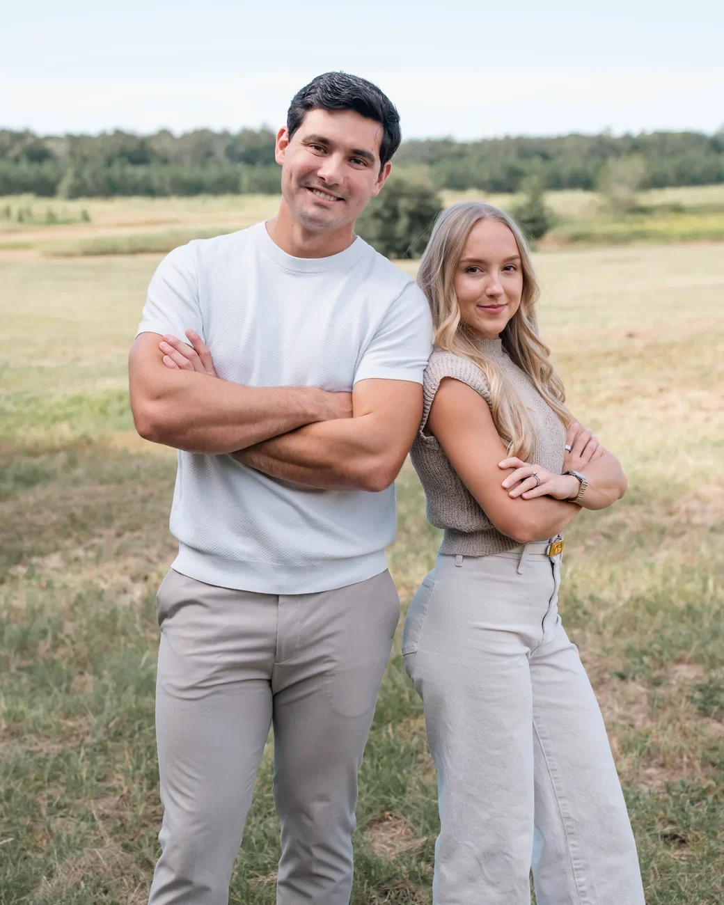 Derrick and Emily Sumner stand back to back with arms crossed in a sunny grassy field.