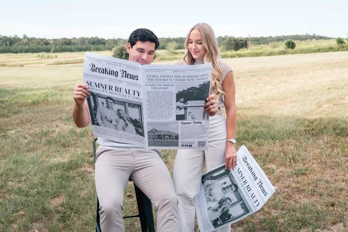 Derrick and Emily Sumner read a Breaking News newspaper announcing Sumner Realty in a grassy field.