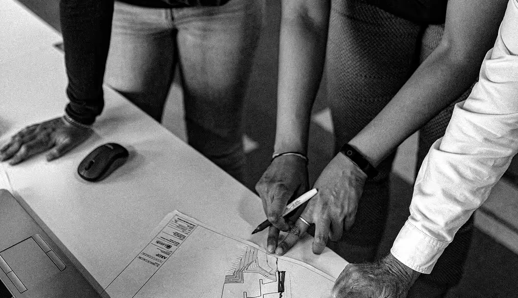 Three people reviewing and pointing at a document with architectural plans on a desk.