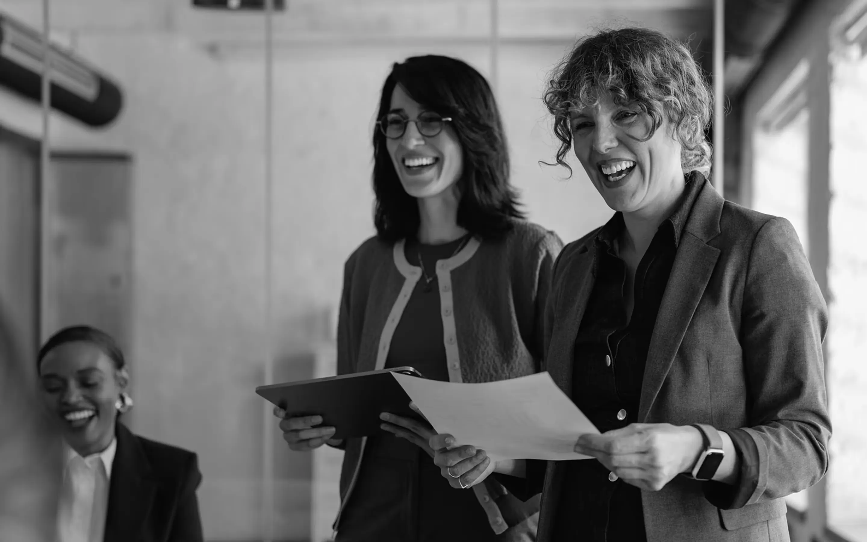 Three businesswomen laughing and reviewing documents in an office setting.