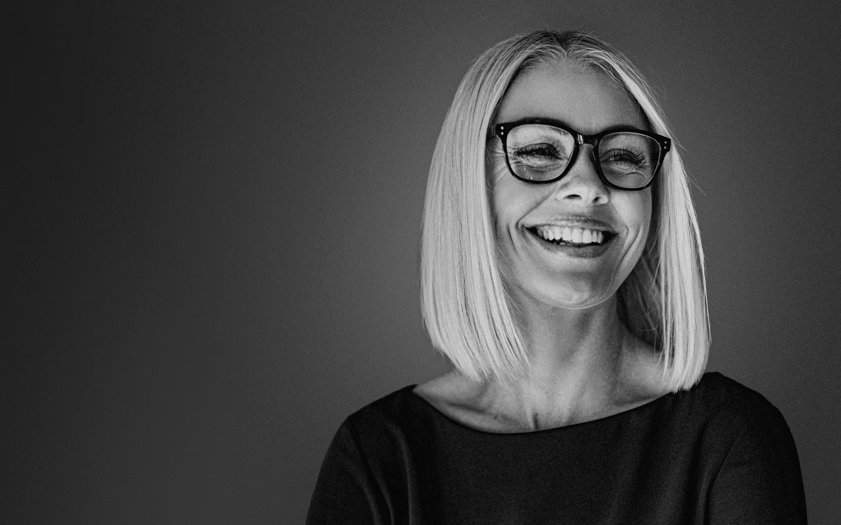 Smiling woman with straight blonde hair and black glasses wearing a dark top, against a plain dark background.