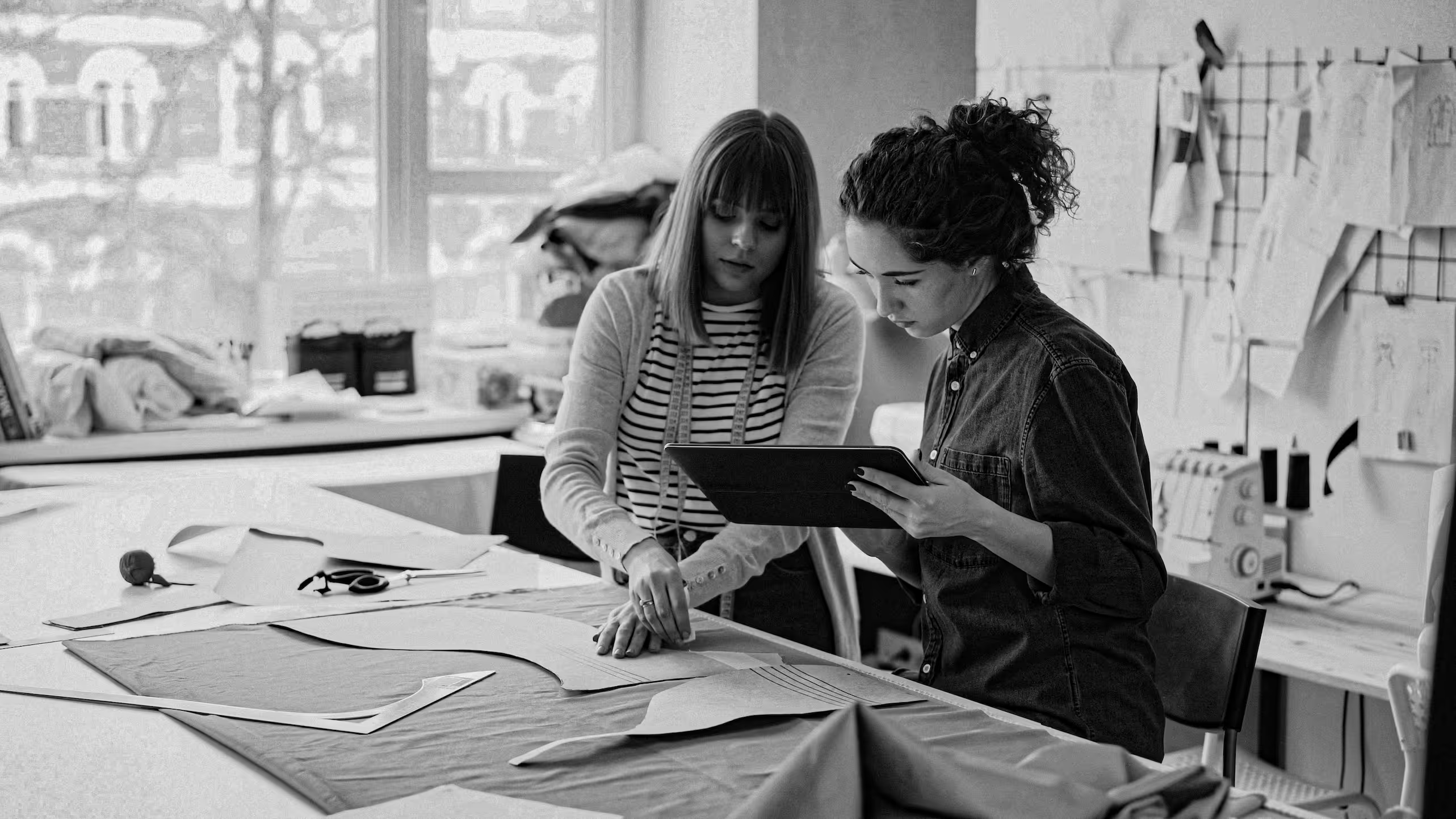 Two women working on fabric patterns at a table in a sewing studio with sketches and tools nearby.