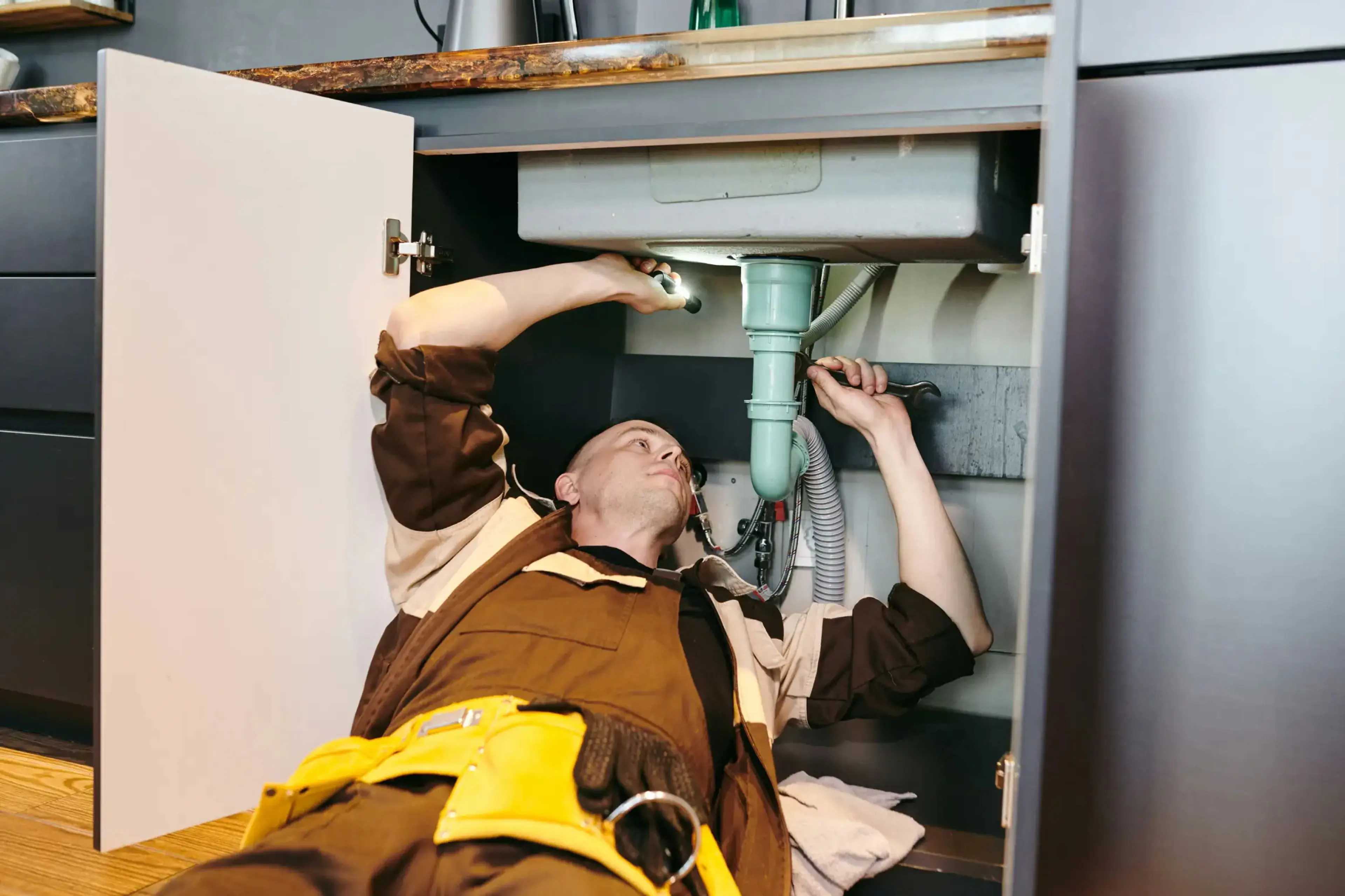 Plumber lying under a kitchen sink repairing the drain pipe with a wrench