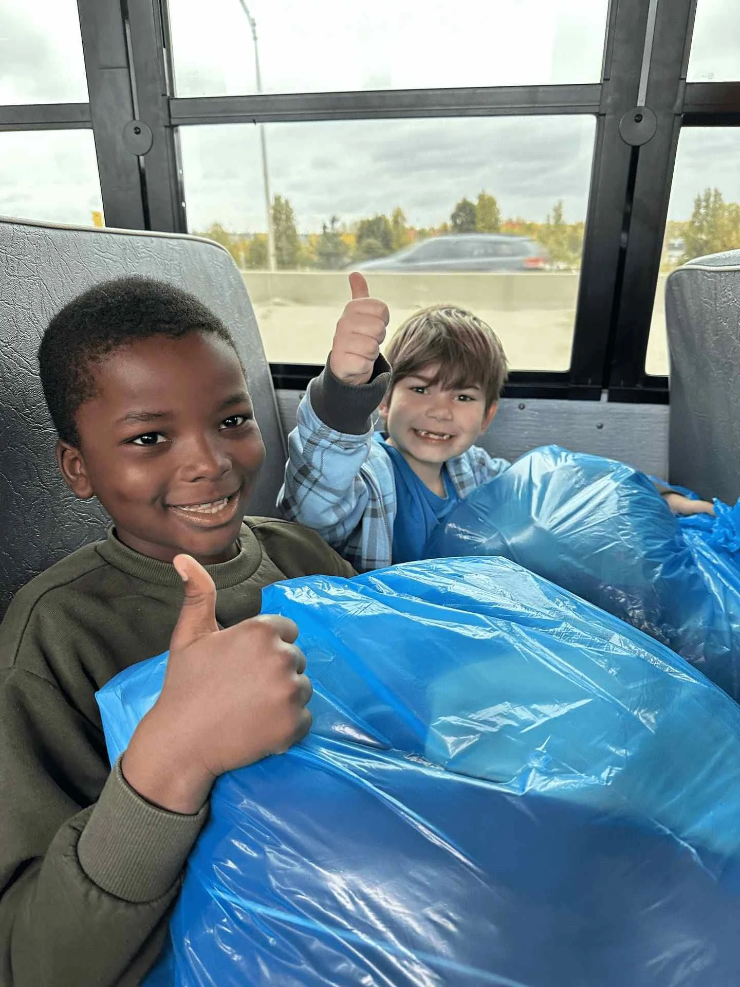 Deux enfants souriants, pouces levés, dans l'autobus avec leurs sacs de vêtements neufs du Club Kiwanis de Québec