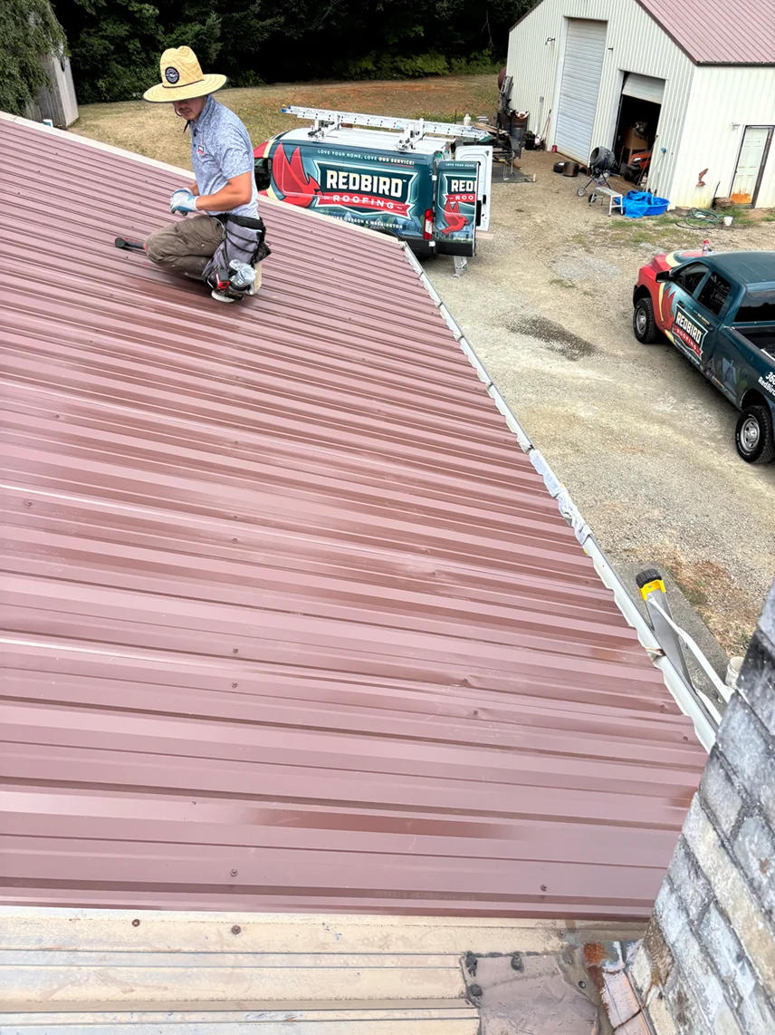 RedBird Roofing crew member fastening panels on a brown metal roof beside a RedBird Roofing van
