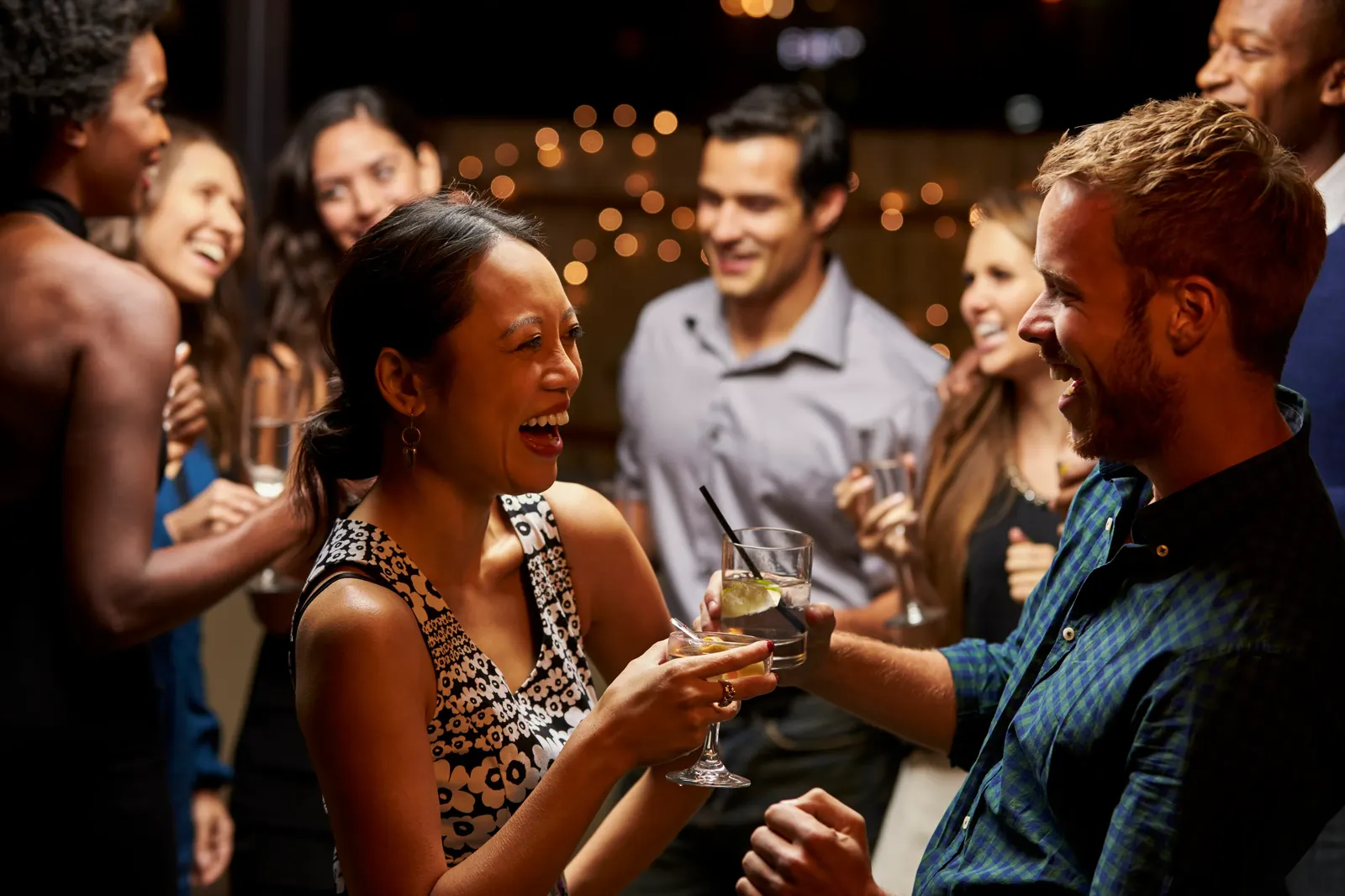 Two friends laughing and clinking drinks at a crowded evening party