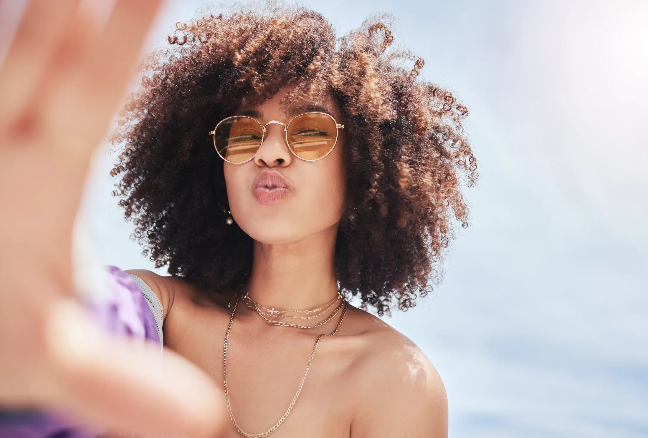 Woman with curly hair and gold sunglasses blowing a kiss at the camera while taking a selfie