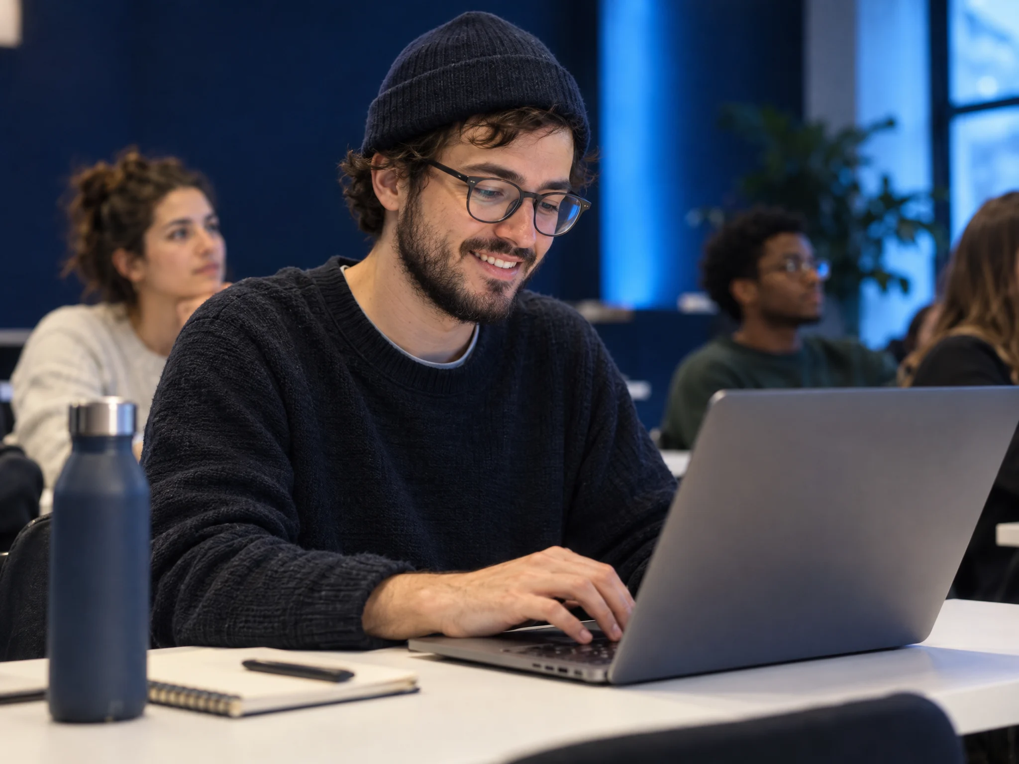 A freelancer in glasses and a beanie typing on a laptop in class
