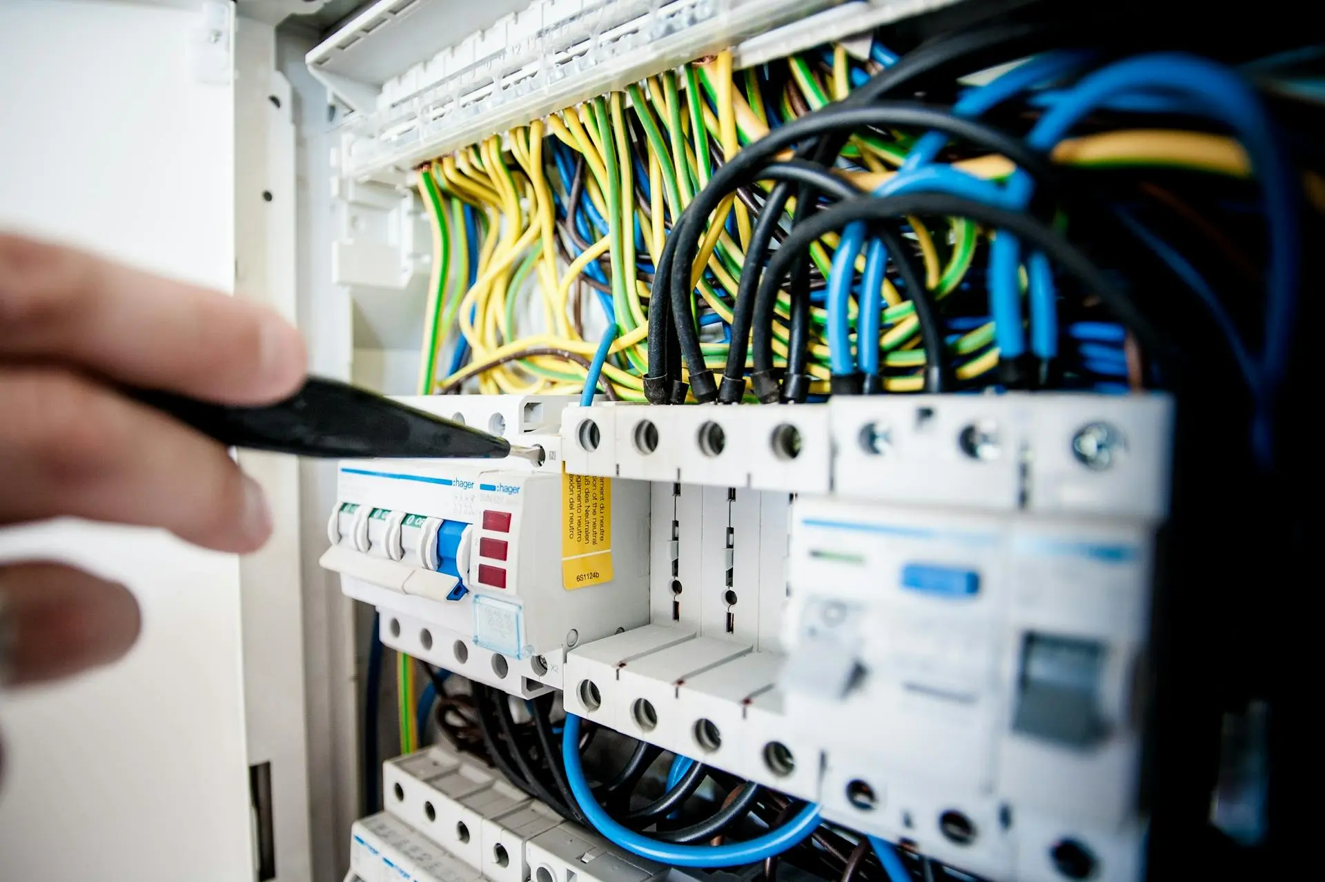Technician pointing a screwdriver at a circuit breaker in an open electrical panel full of wiring.