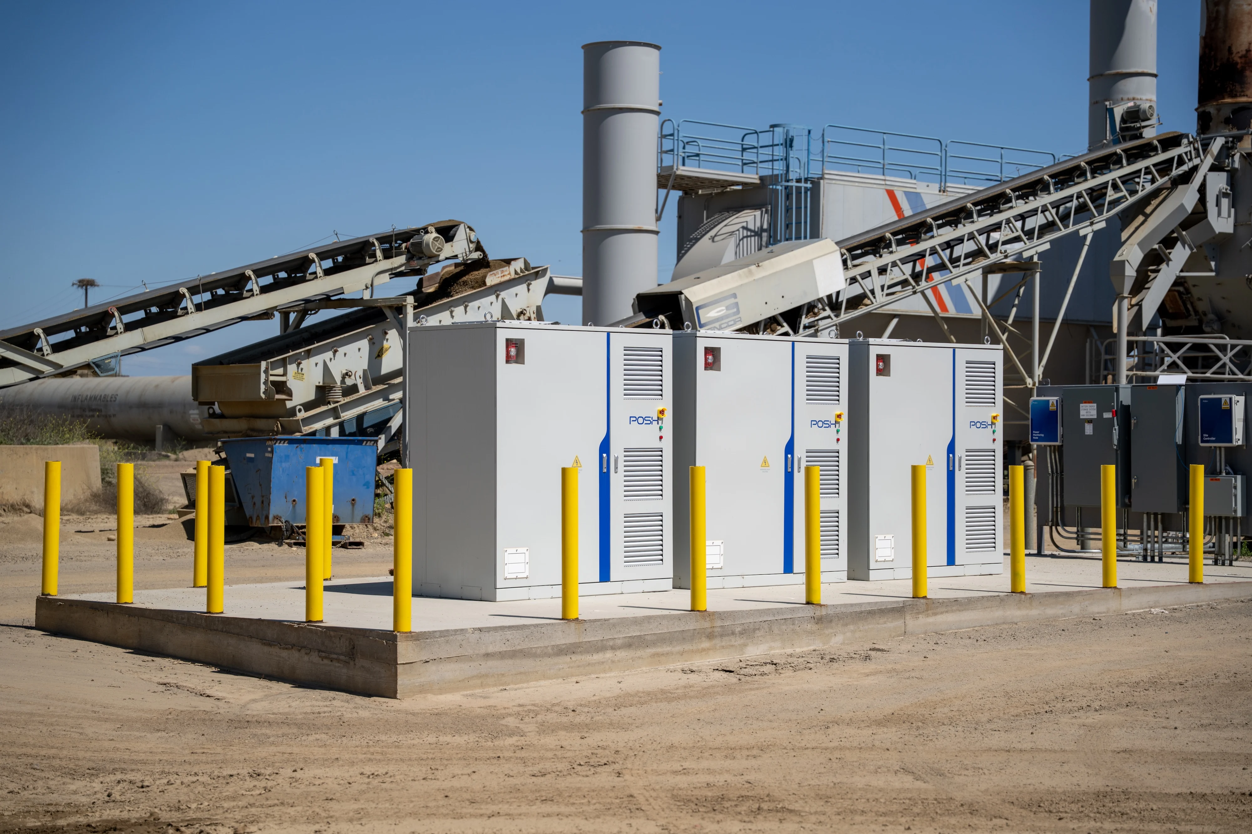 Three POSH Station battery cabinets on a concrete pad protected by yellow bollards at a Teichert industrial plant, with conveyors and silos behind.