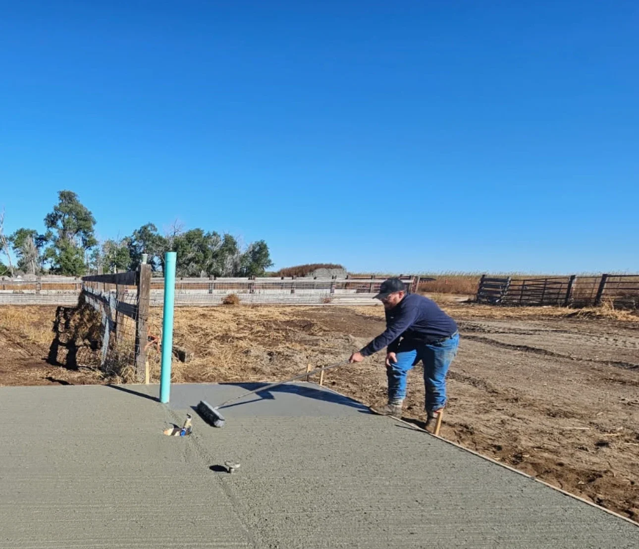Kuntz Pump & Well Supply crew member broom-finishing a fresh concrete pad around a well riser at a ranch corral