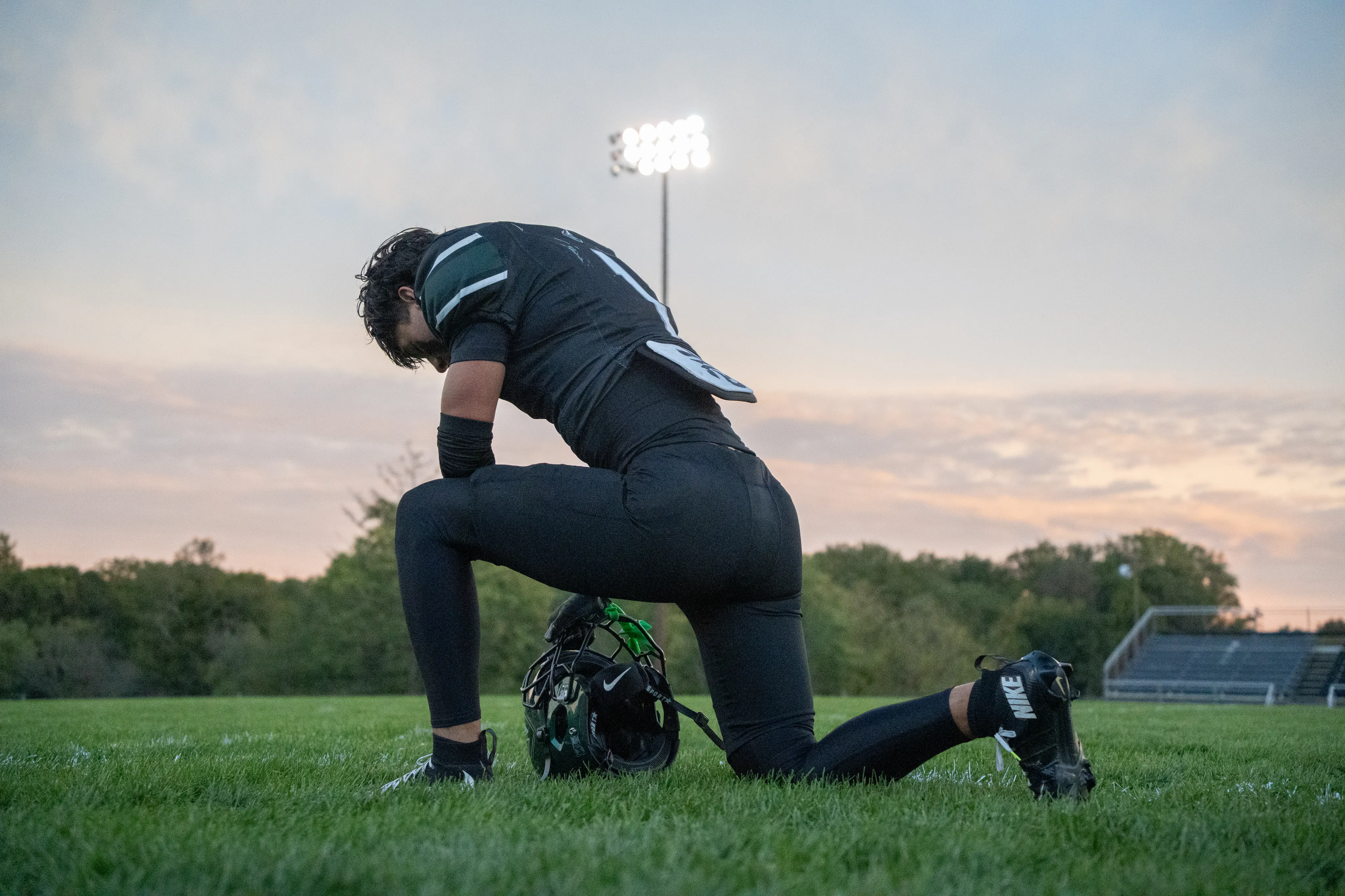 Football player kneeling beside a helmet on the field at dusk