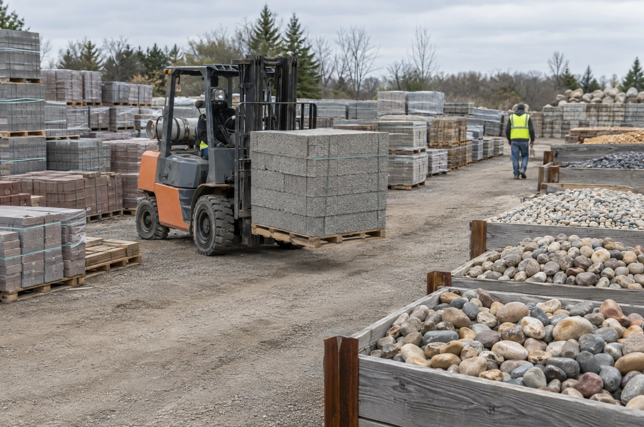 Forklift moving a pallet of retaining wall block past paver pallets and bins of river rock in a hardscape yard