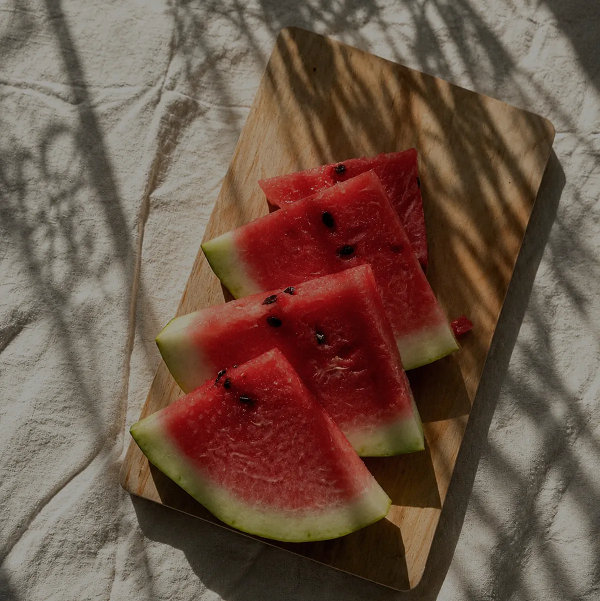 Watermelon slices on a wooden board, with leaf shadows across linen.