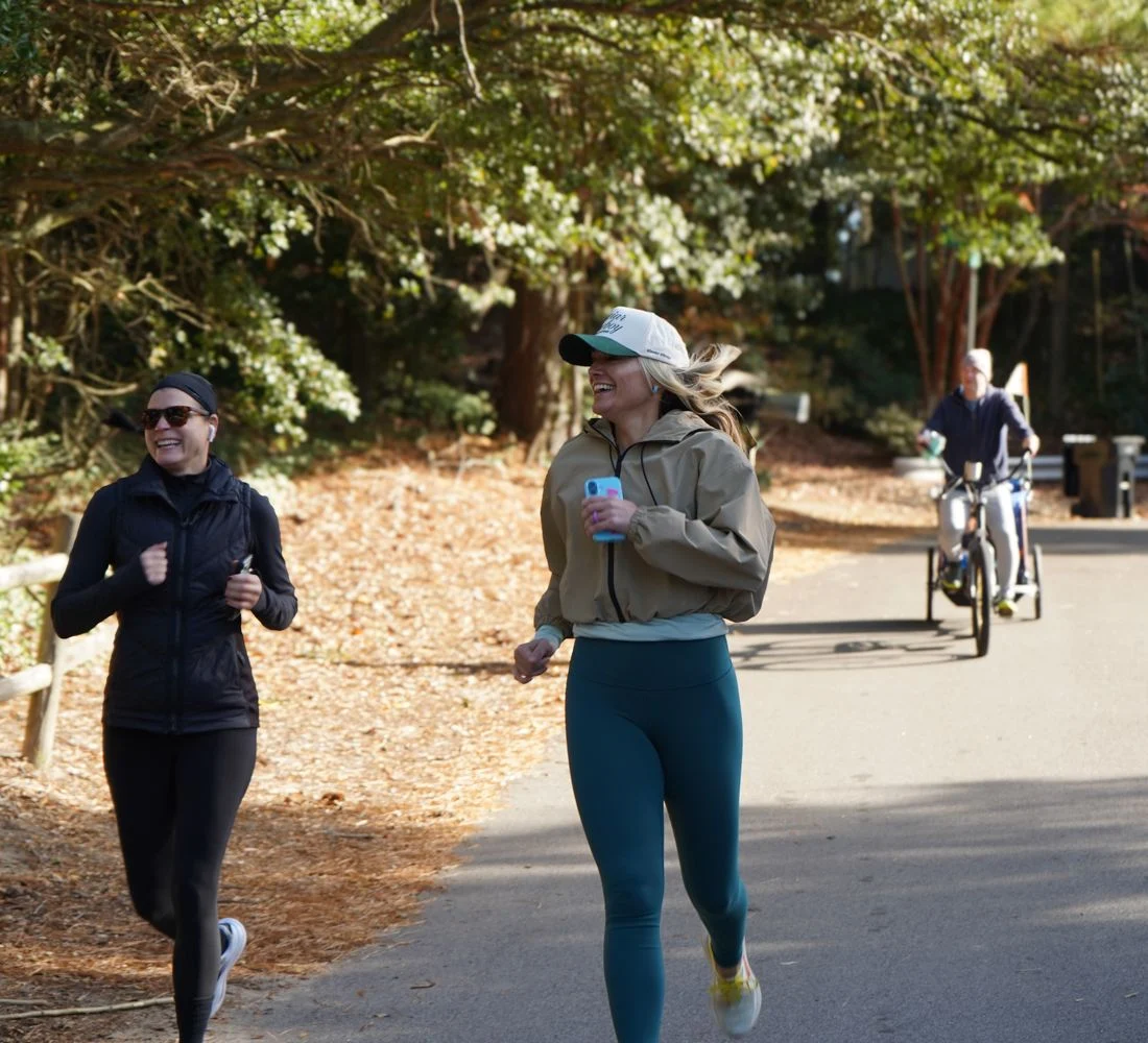 Two runners smile as they jog down the paved path, with a person on an adaptive trike behind them.