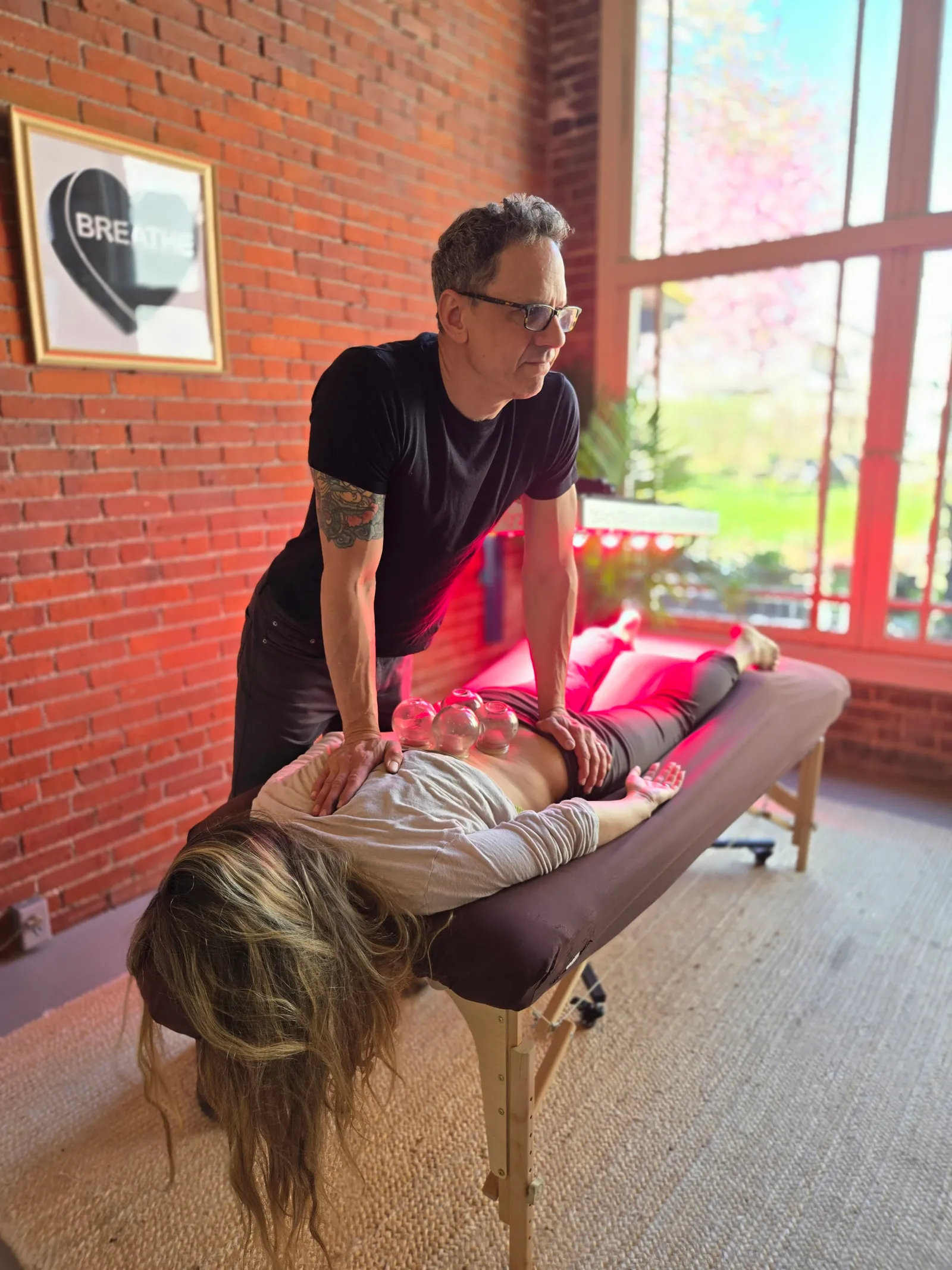 Jason Rinker working with his hands on a patient's back beside glass cups, in his brick-walled room by the garden window