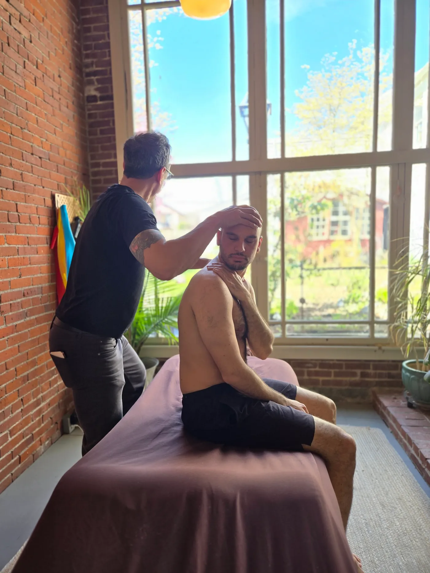 Jason Rinker assessing a seated patient's neck and shoulder in front of the garden window