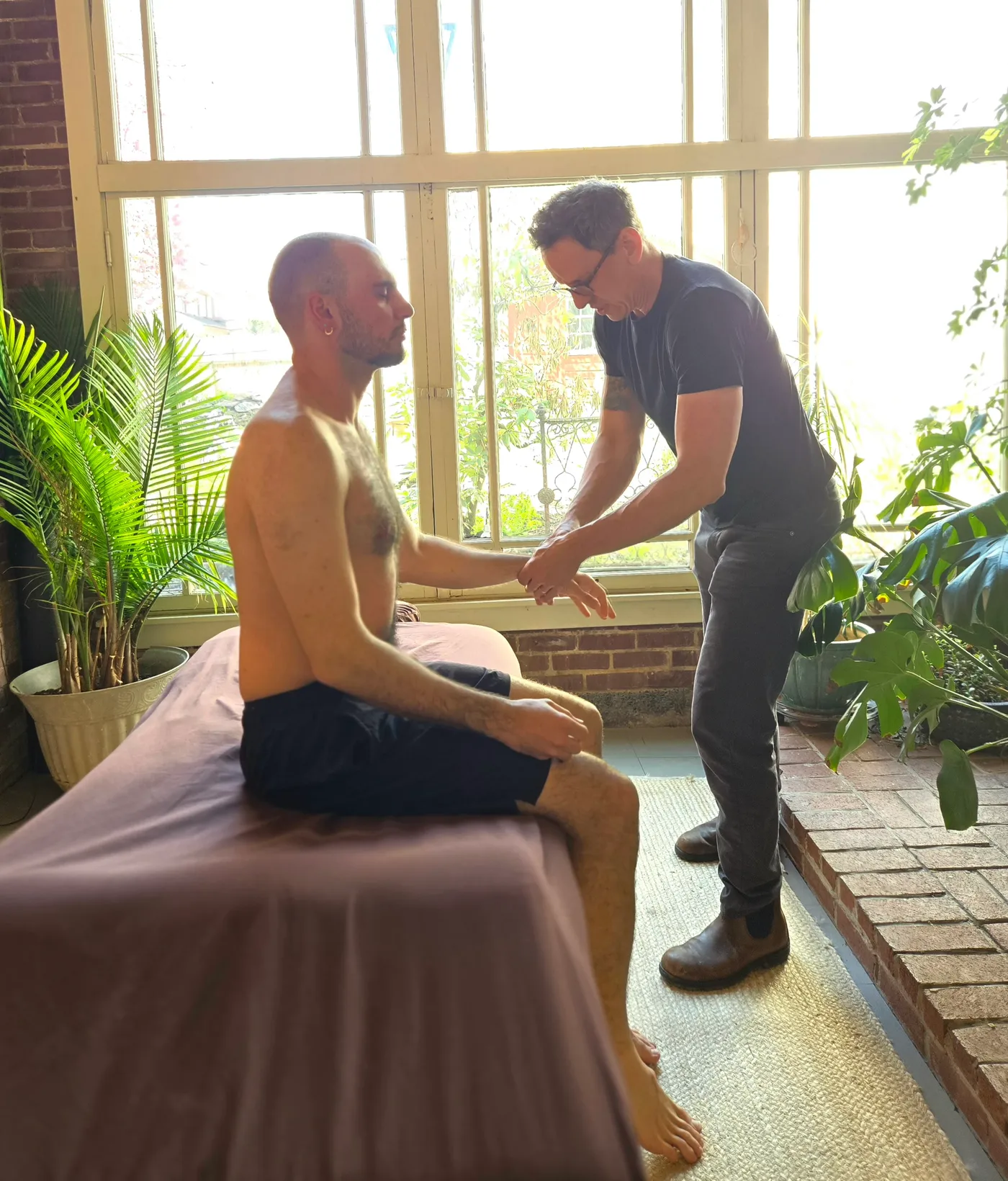 Jason Rinker assessing a seated male patient's wrist in the sunlit treatment room
