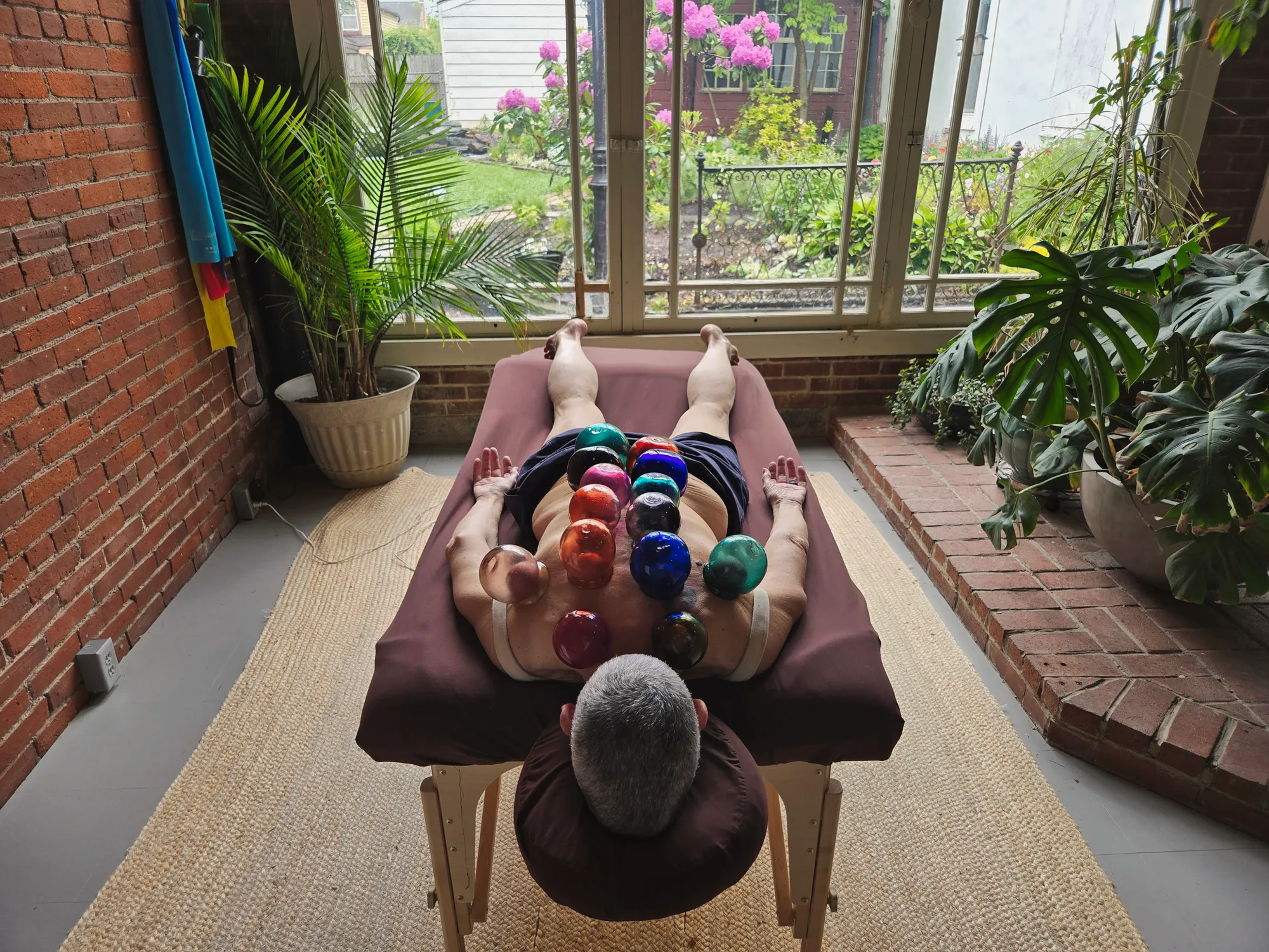 A patient resting face down with colored glass cups along his back, in the treatment room overlooking the garden