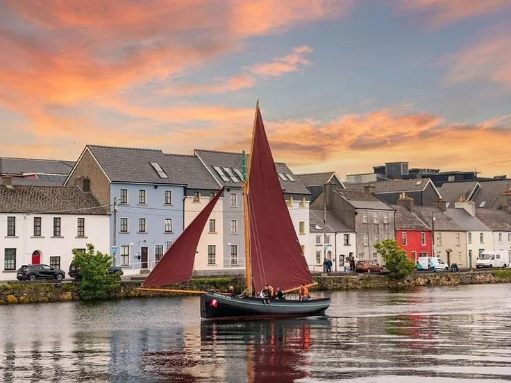 A Galway hooker with red sails passing the Long Walk in Galway at sunset