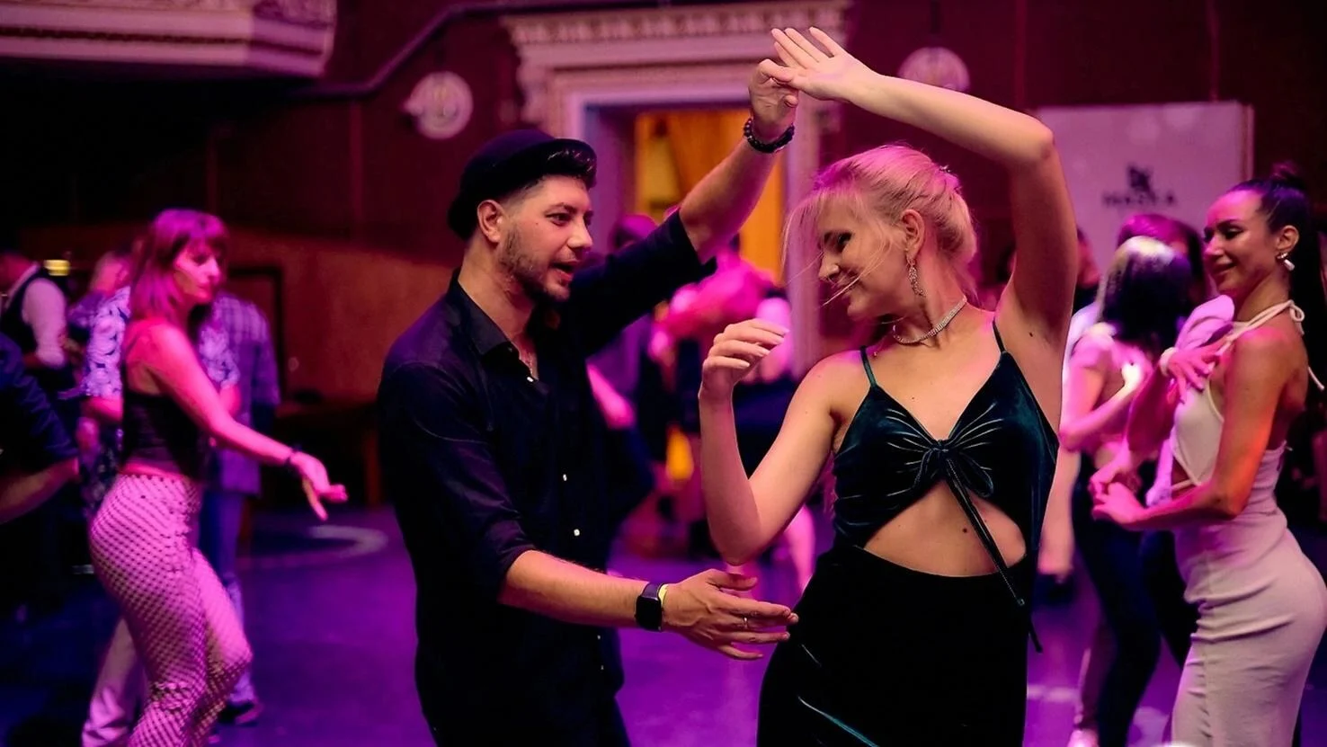 A salsa couple mid-turn on a crowded dance floor under pink lights