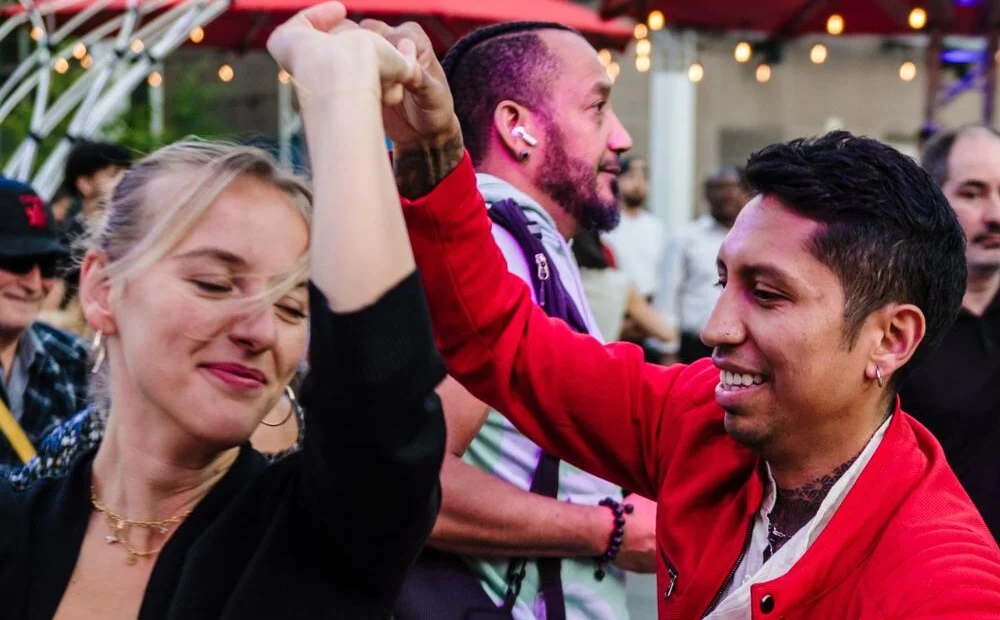 A smiling couple dances salsa outdoors surrounded by the crowd